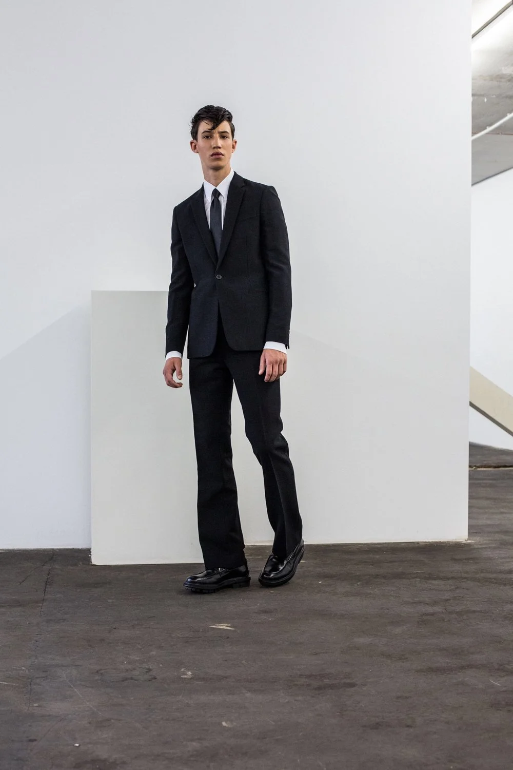 Young man in black suit and tie standing indoors on dark floor with white wall background.