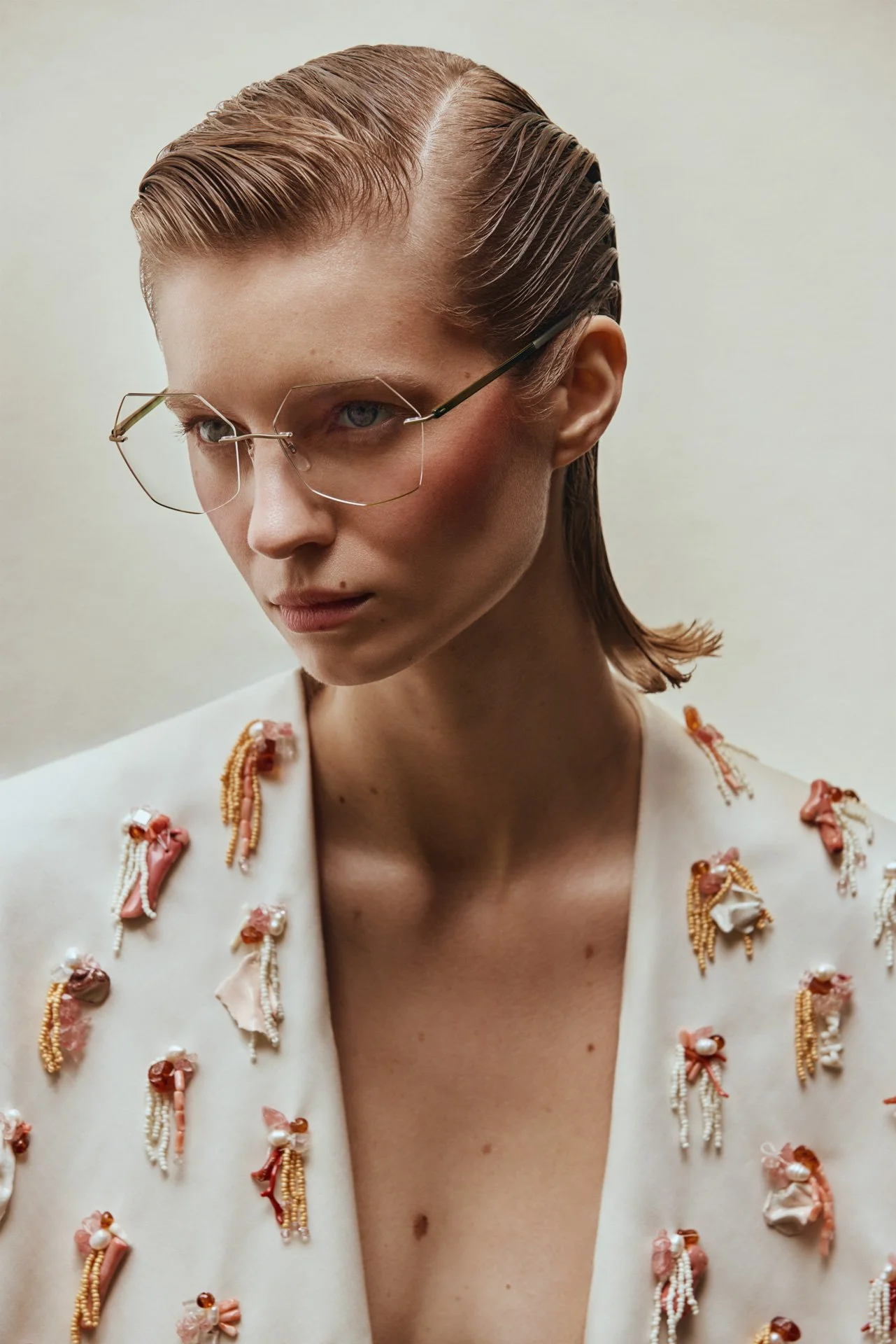 A woman with slicked-back hair, wearing large fashionable glasses and a white jacket decorated with pink and white fabric embellishments.