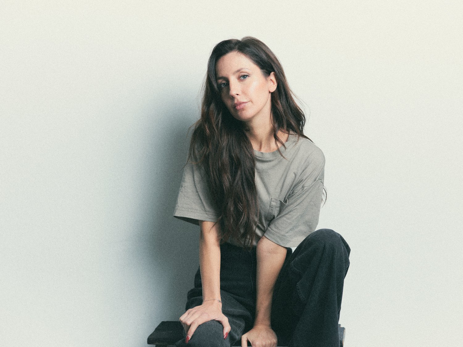 A young woman with long wavy brown hair, wearing a loose grey t-shirt and black pants, sitting with one leg bent and her hand resting on her knee against a plain light-colored wall.