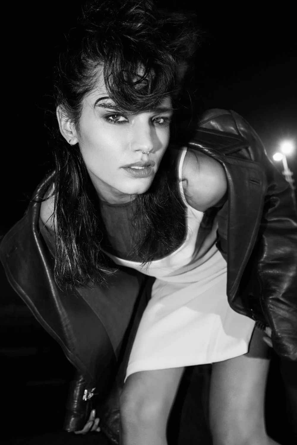 Black and white photo of a woman with dark hair, wearing a leather jacket and a white dress, looking at the camera with a confident expression.