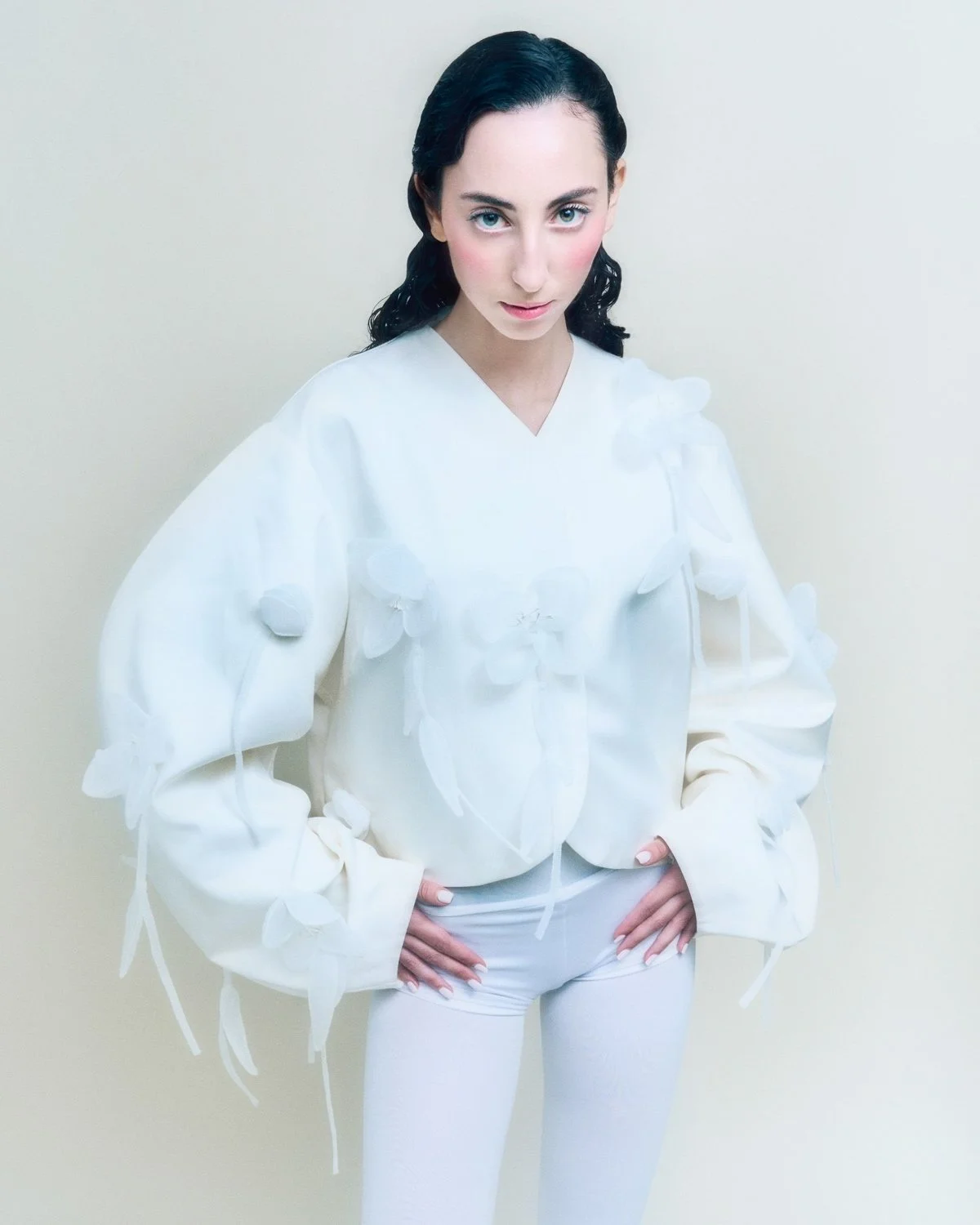 Woman with dark hair in a white outfit with floral details, standing against a plain wall.
