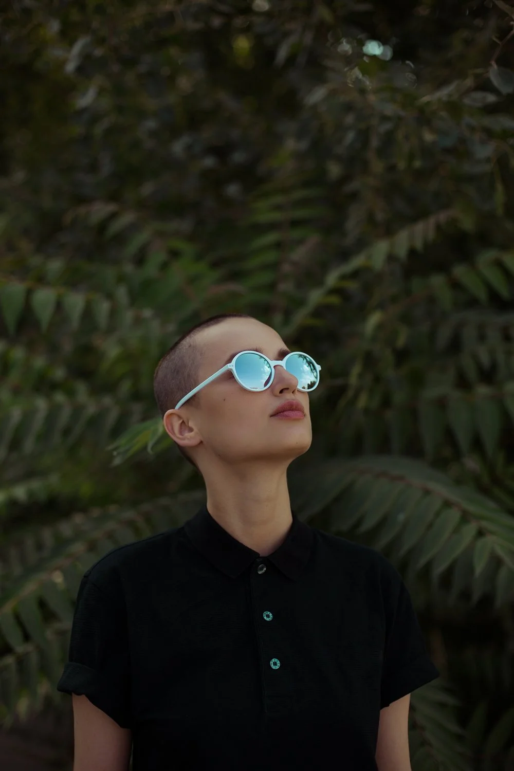 A woman wearing round sunglasses with reflective blue lenses, a black collared shirt, and short buzzed hair, standing outdoors in front of dense green foliage.