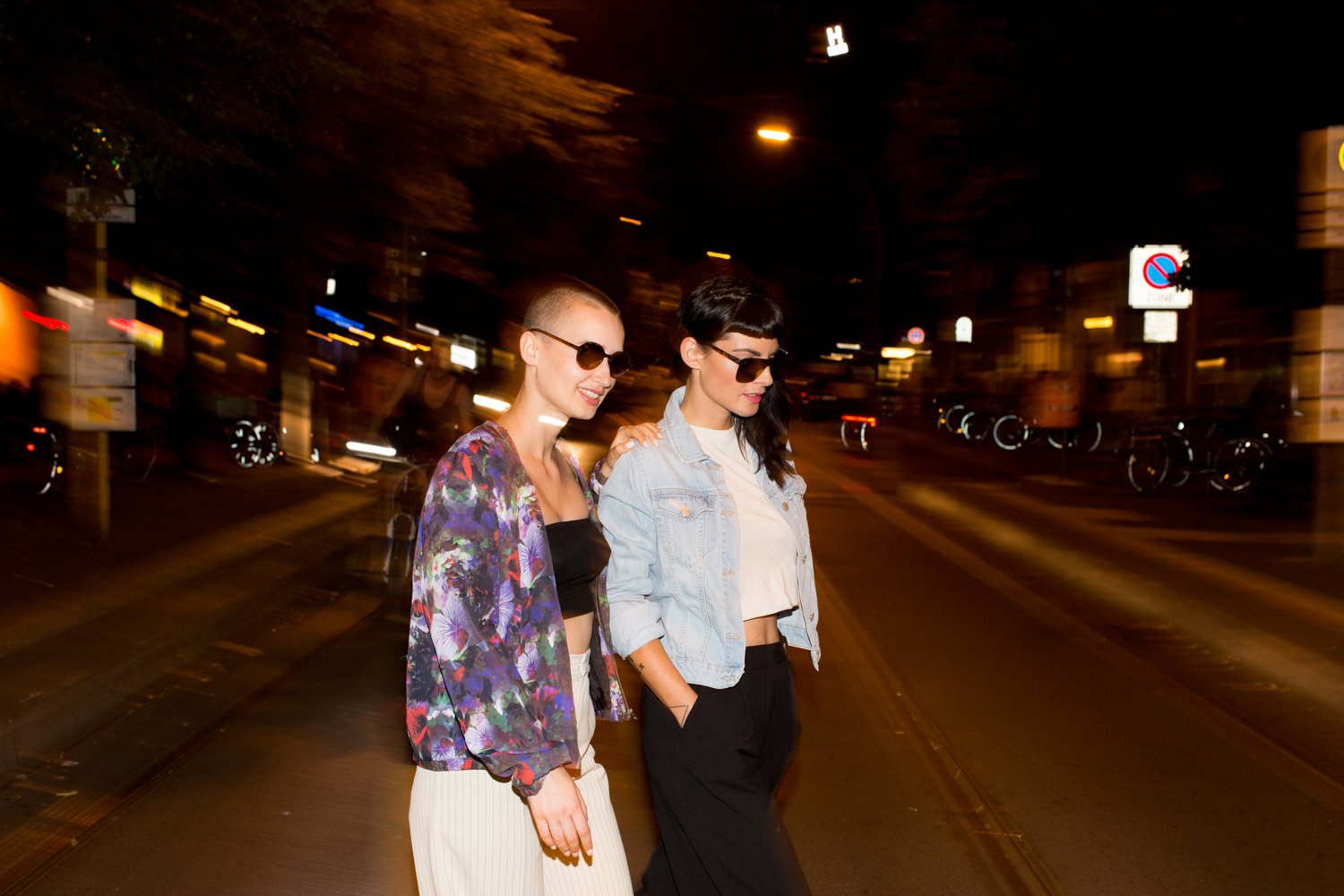 Two women walking on a city street at night, wearing sunglasses and casual clothes, with blurred lights in the background.