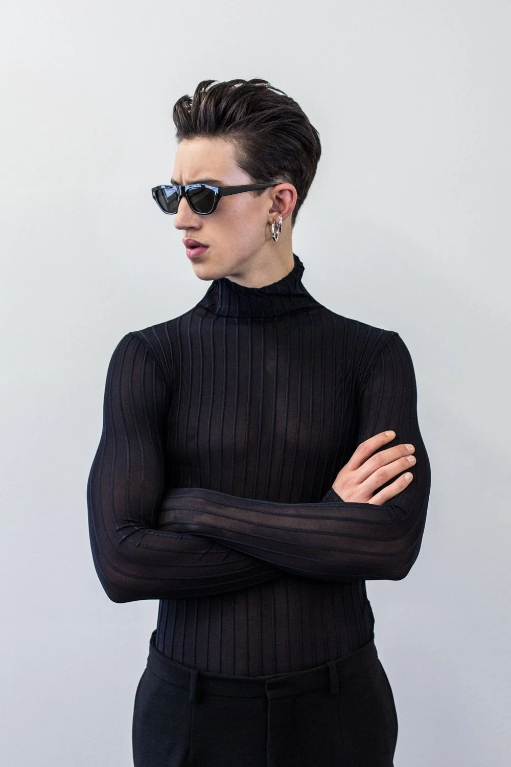 Person with short dark hair wearing sunglasses, earrings, and a black, long-sleeved turtleneck, crossing arms against a plain light background.