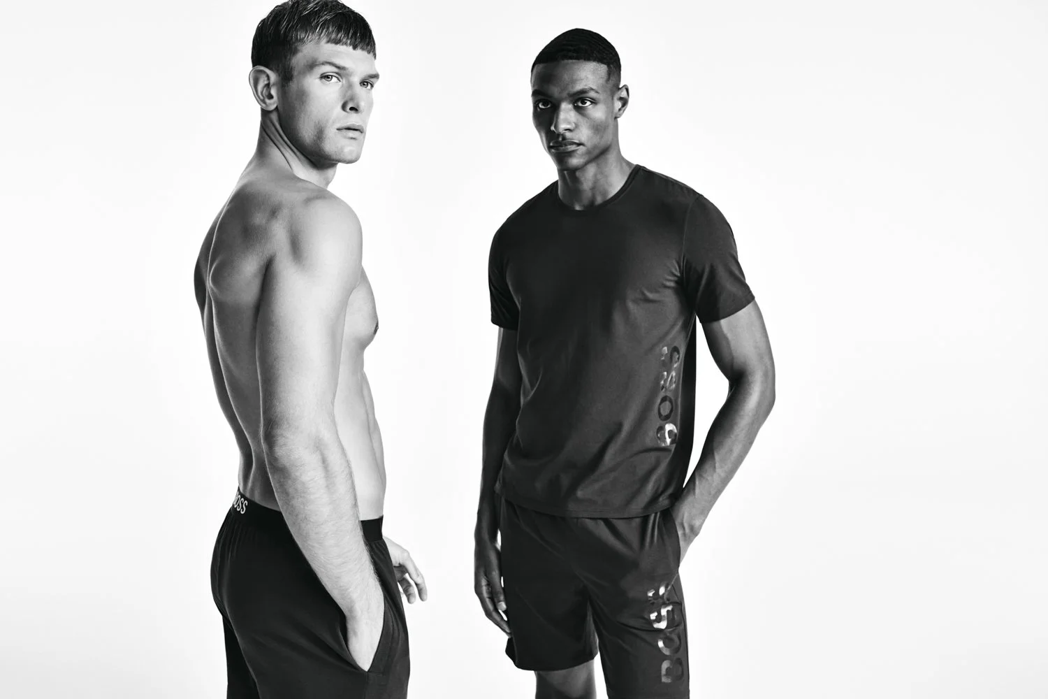 Black and white photo of two young men standing against a plain background. One shirtless man with a buzz cut faces the camera in a three-quarter view, with his hands in his shorts pockets. The other man wears a T-shirt and shorts, both with the BOSS