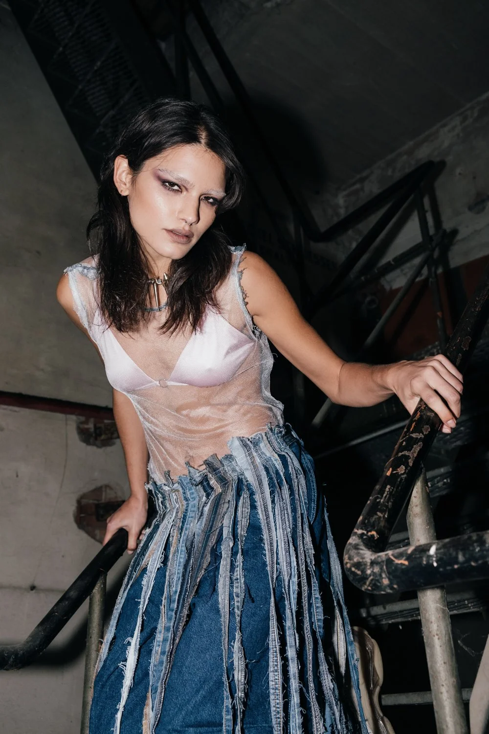 A woman with dark hair, wearing a sheer tank top and a denim skirt with torn and striped fabric, standing on a worn metal staircase in an industrial setting with concrete walls.