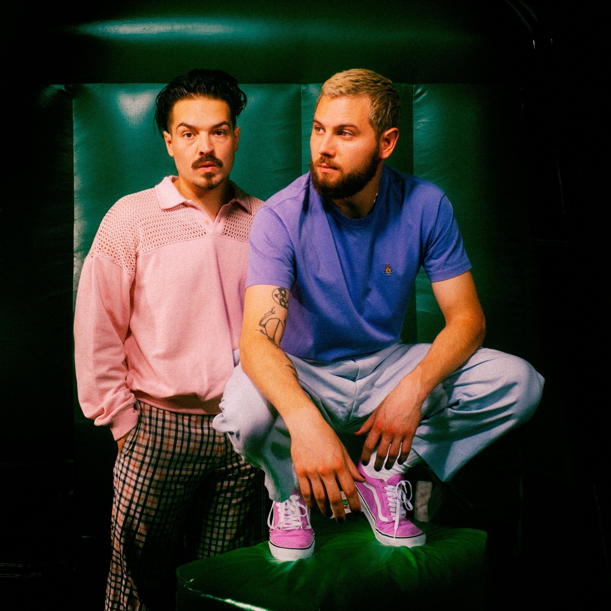Two young men posing against a green background, one standing with hands in pockets wearing a pink shirt and checkered pants, and one sitting on a stool with one leg up wearing a blue t-shirt, white pants, and pink sneakers.