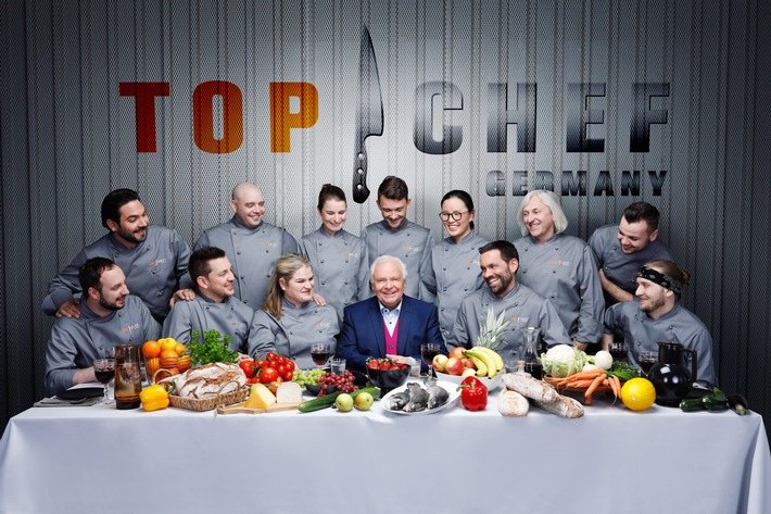 Group of chefs and a man in a suit sitting at a table with various fruits, vegetables, and cooking ingredients in front of a "Top Chef Germany" sign.