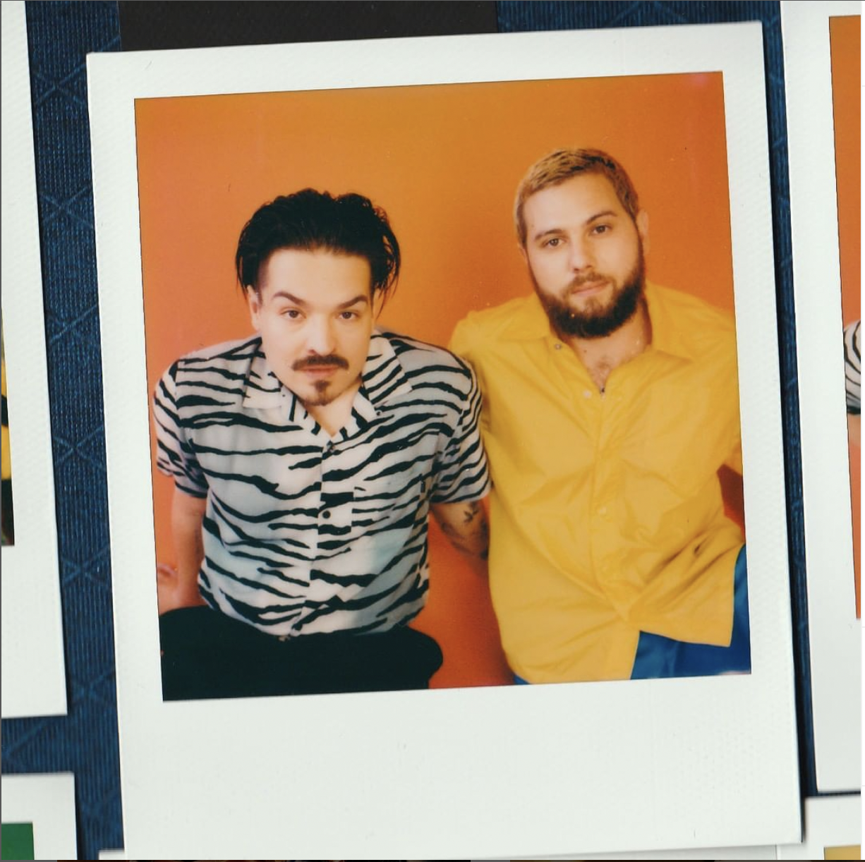 Two men sitting against an orange background. The man on the left wears a zebra-striped shirt and has black hair styled back, with a mustache and goatee. The man on the right wears a yellow button-up shirt, has short, light brown hair, a beard, and a