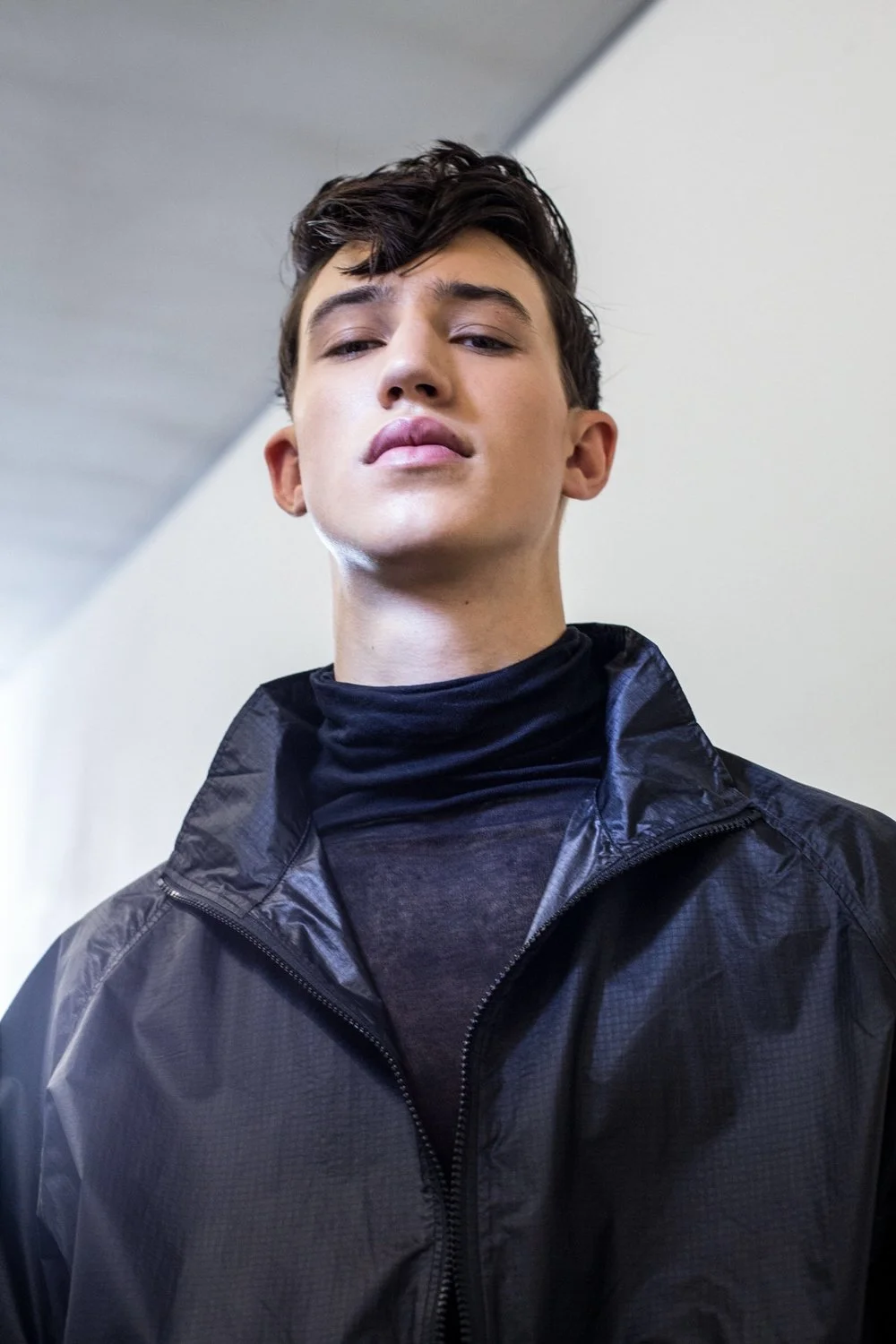 A young man with dark, wavy hair and light skin, wearing a black high-neck turtleneck and a black zip-up jacket, looking slightly upward with a confident expression.