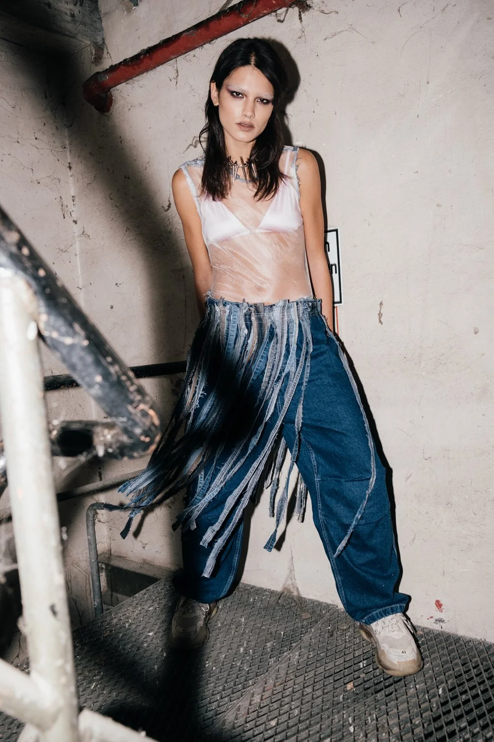 A woman with dark hair and makeup, wearing a sheer sleeveless top, wide-leg distressed jeans with fringe details, and sneakers, standing in a gritty industrial setting against a concrete wall with pipes.