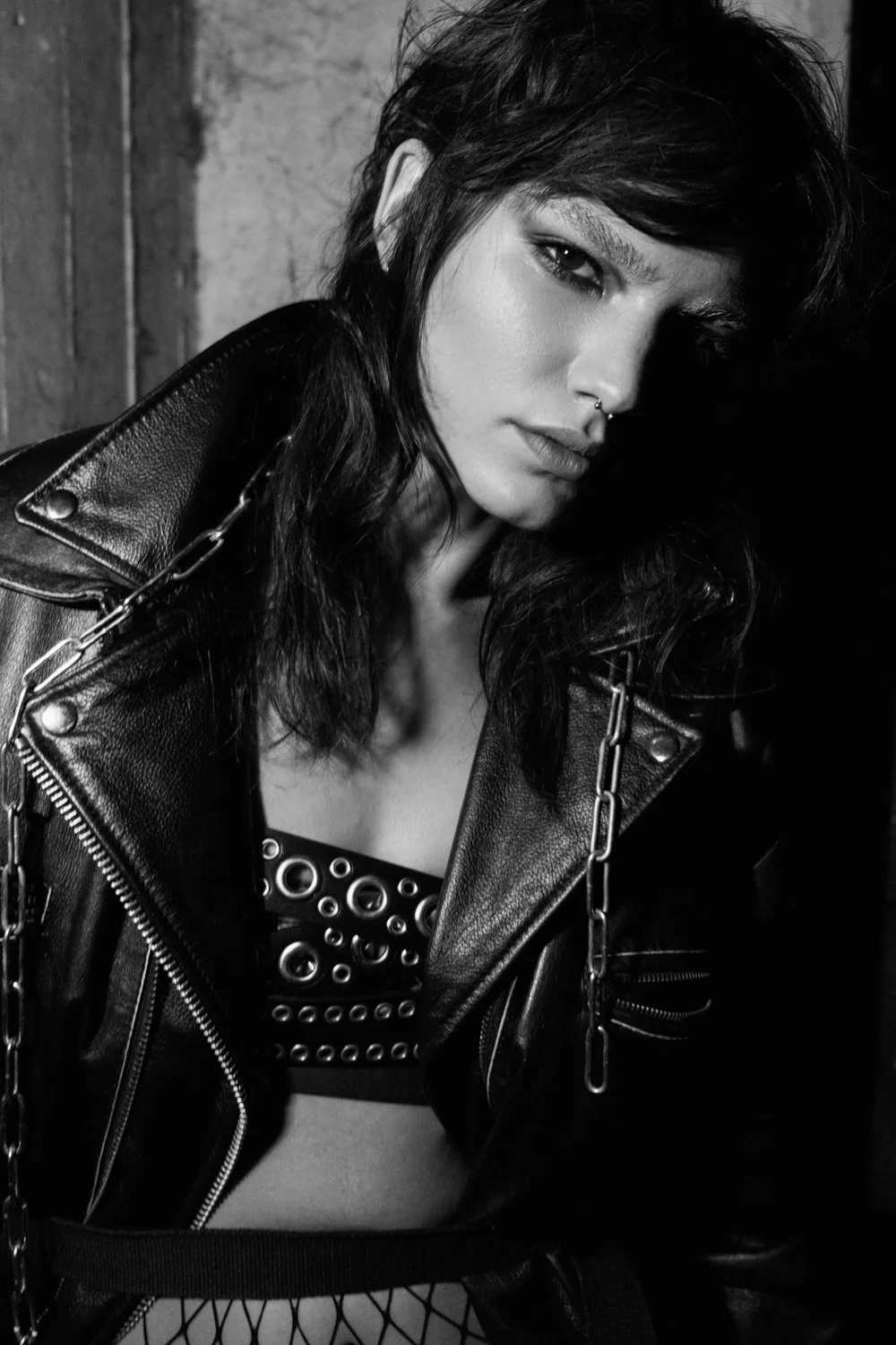 A woman with dark hair styled in waves, wearing a studded top, a leather jacket with chain details, and fishnet stockings, posing against a textured wall.