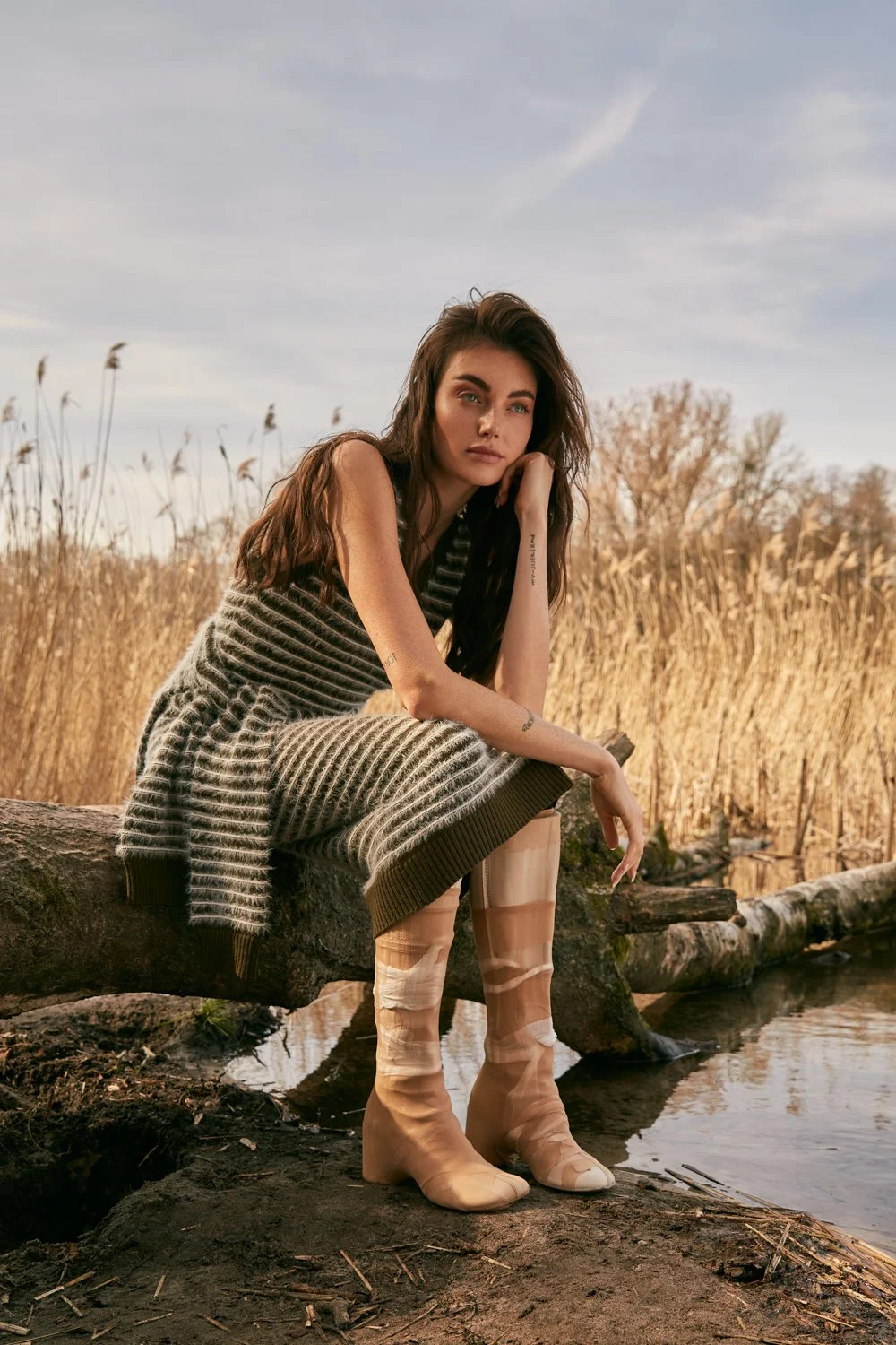 A young woman with long brown hair and tattoos, sitting on a log by a small body of water in a natural outdoor setting with tall dry grass and trees in the background, wearing a striped sleeveless dress and tan boots.