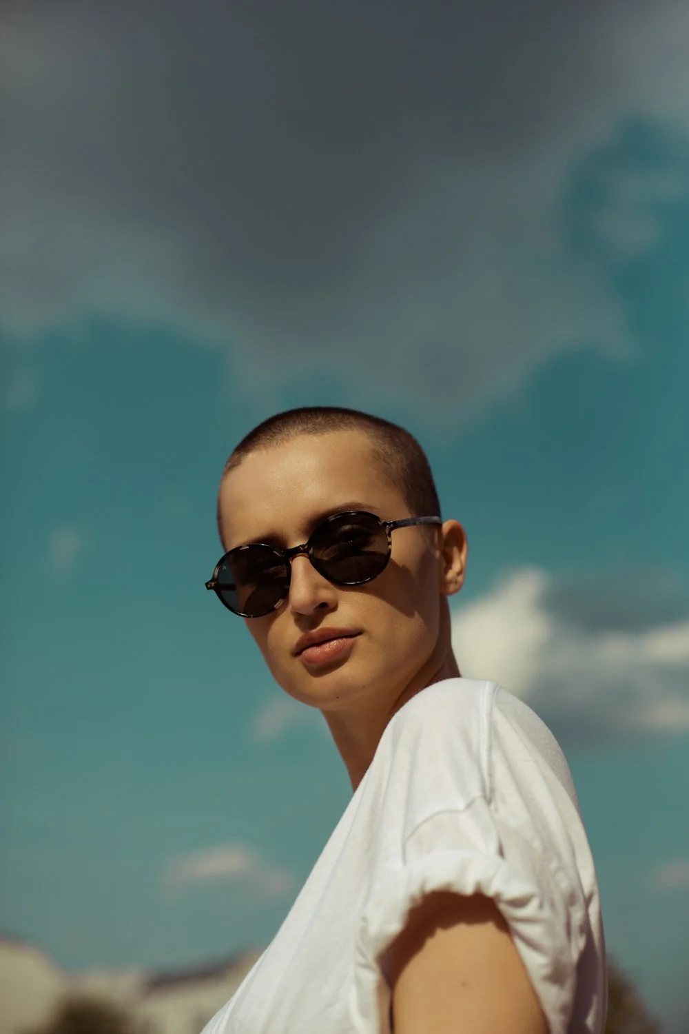 Young woman with short hair wearing sunglasses and a white shirt, outdoors with cloudy sky in background.
