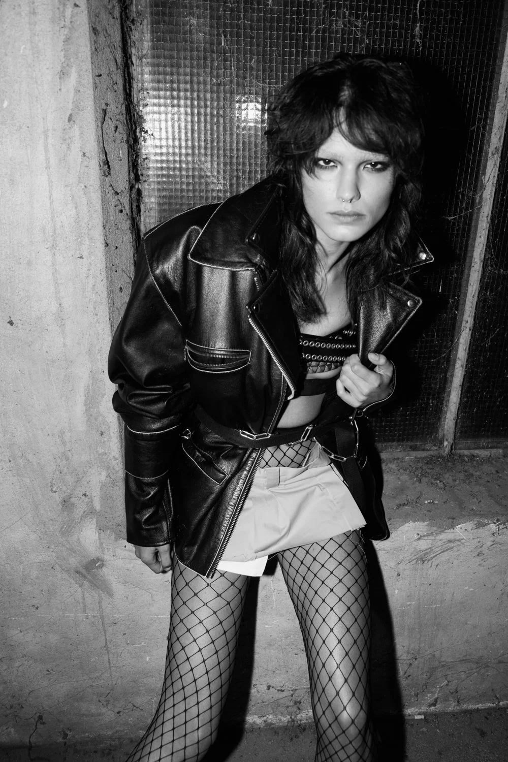 A woman with dark, shaggy hair and a septum piercing, wearing a black leather jacket, fishnet stockings, and a short skirt, standing in an urban alleyway with a gritty, textured wall and metal door behind her, looking directly at the camera.