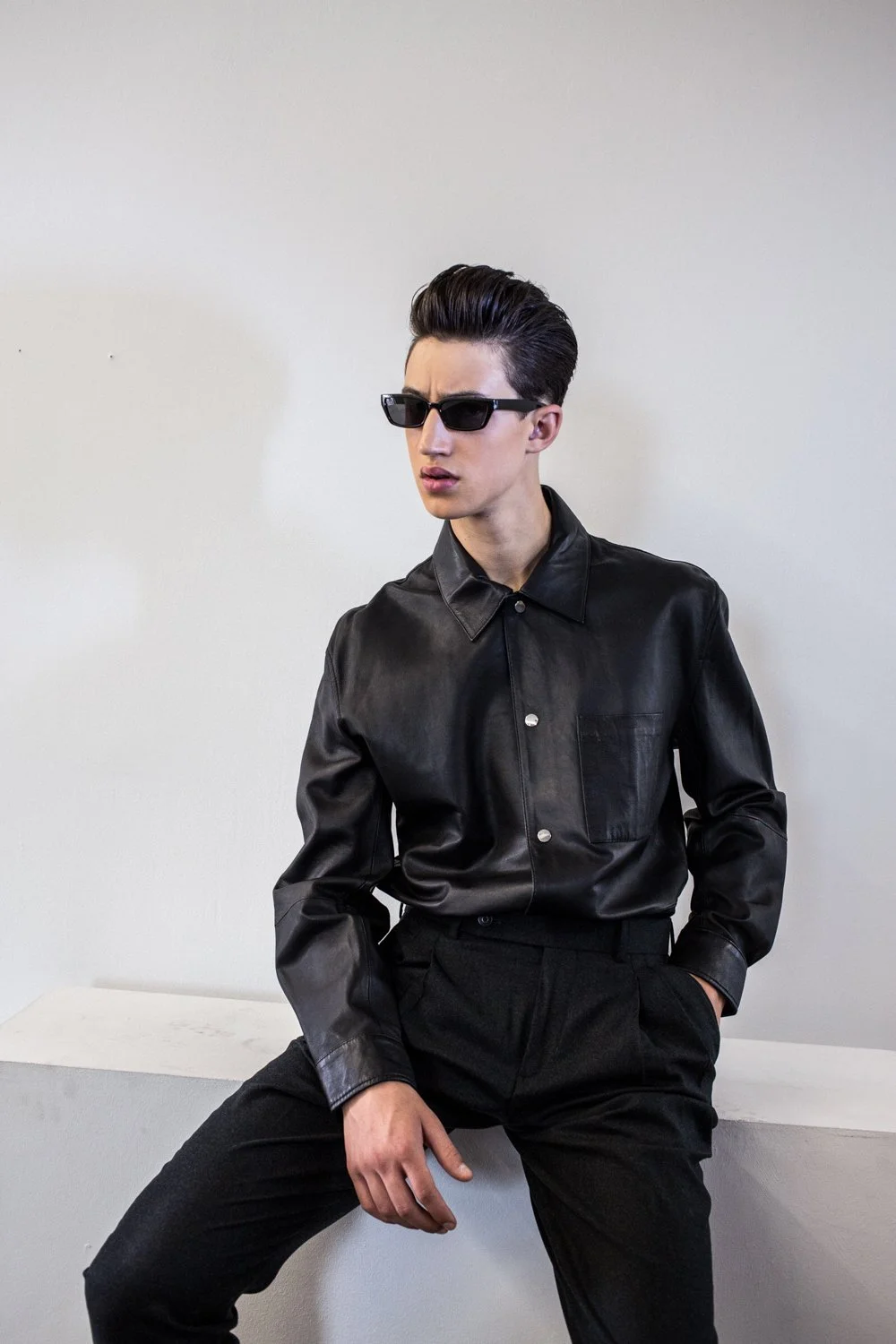 Young man wearing black leather shirt and black pants, sunglasses, leaning against a white wall with one hand on his thigh and the other in his pocket.