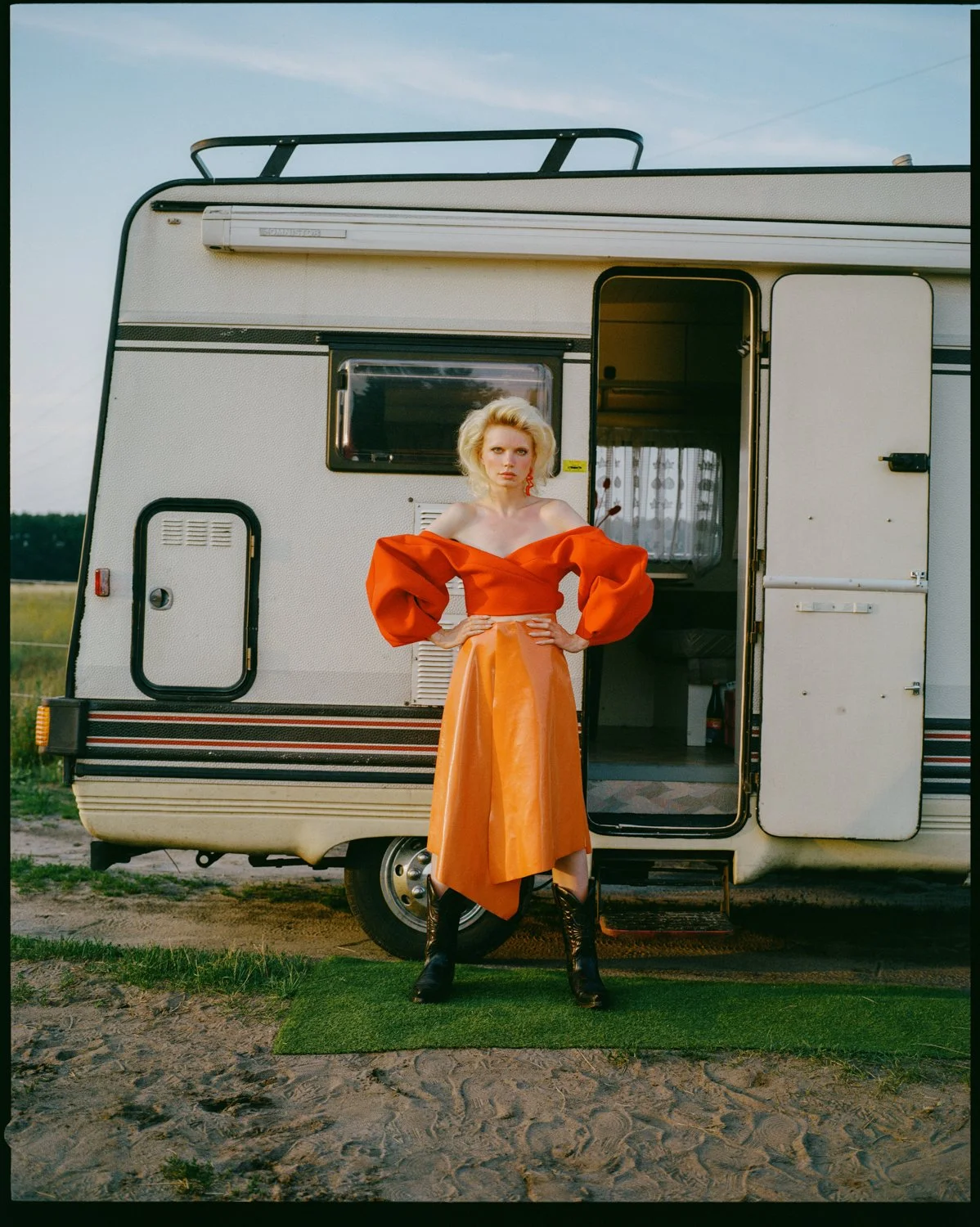 A woman with blonde hair stands with her hands on her hips in front of a white camper, wearing an orange off-the-shoulder top and a matching orange skirt with black boots, outdoors on a grassy area.