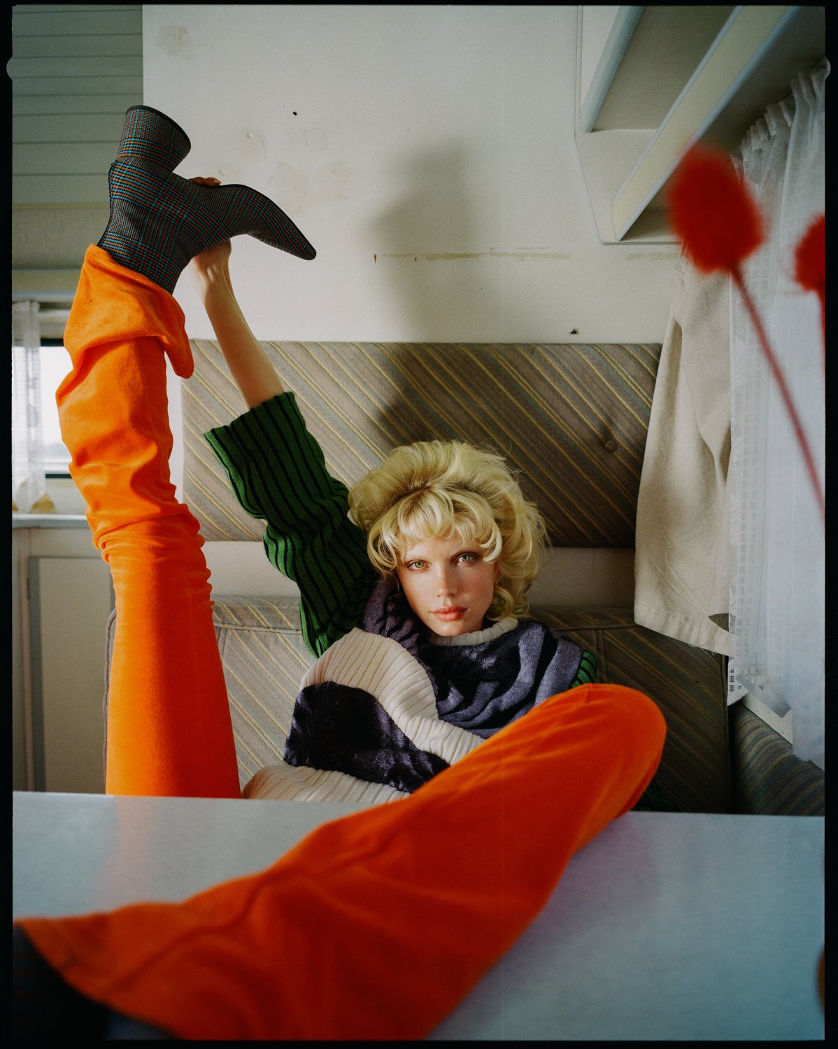 A woman lying on a patterned sofa with her head resting on the armrest, looking at the camera. She has blonde curly hair, and is wearing a multicolored sweater. Her legs are raised, with one in orange pants and a checked boot. The scene appears to be