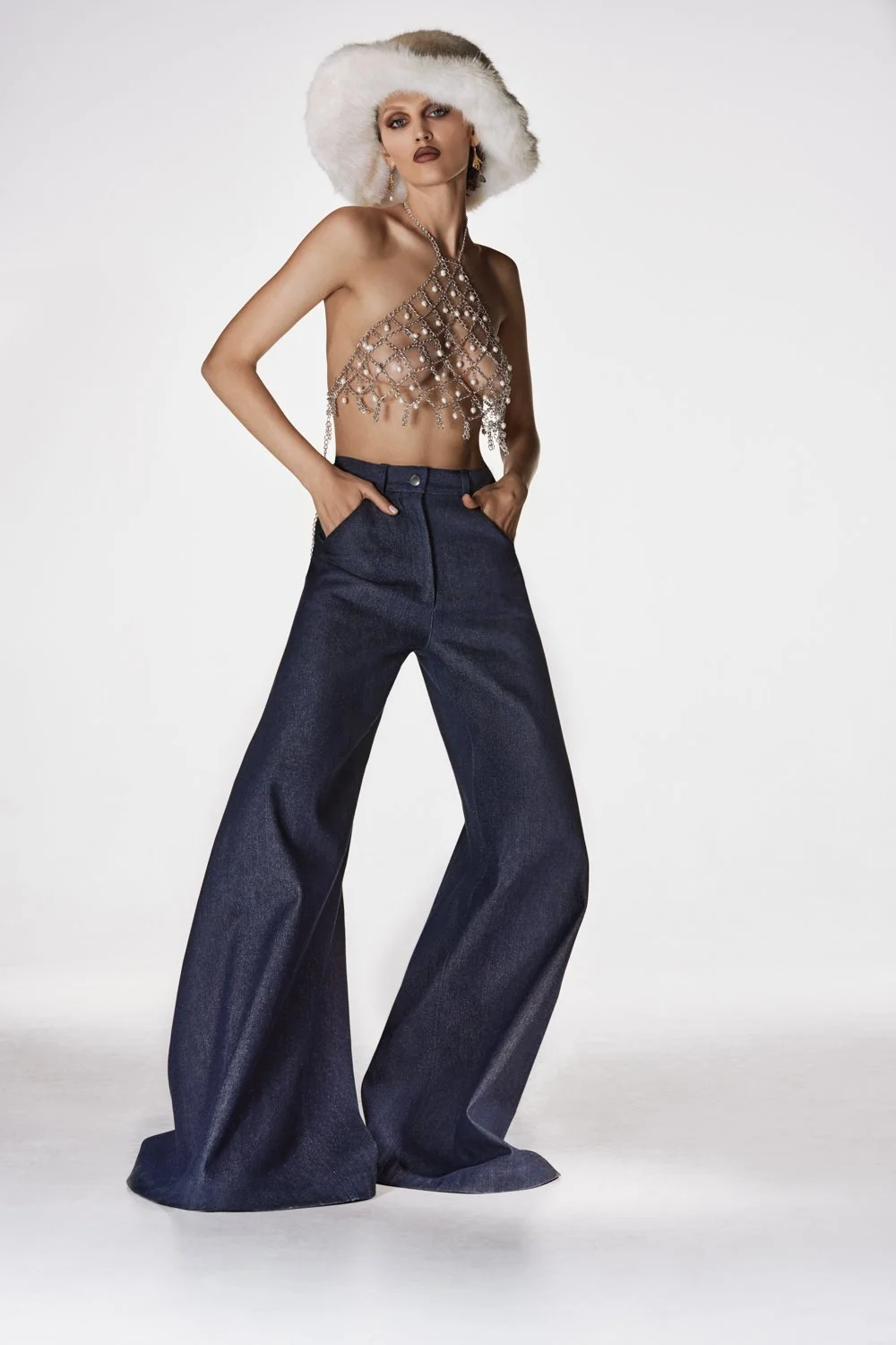 Model wearing high-waisted wide-leg jeans, a chainmail top, statement earrings, and a fluffy white hat, standing against a plain white background.