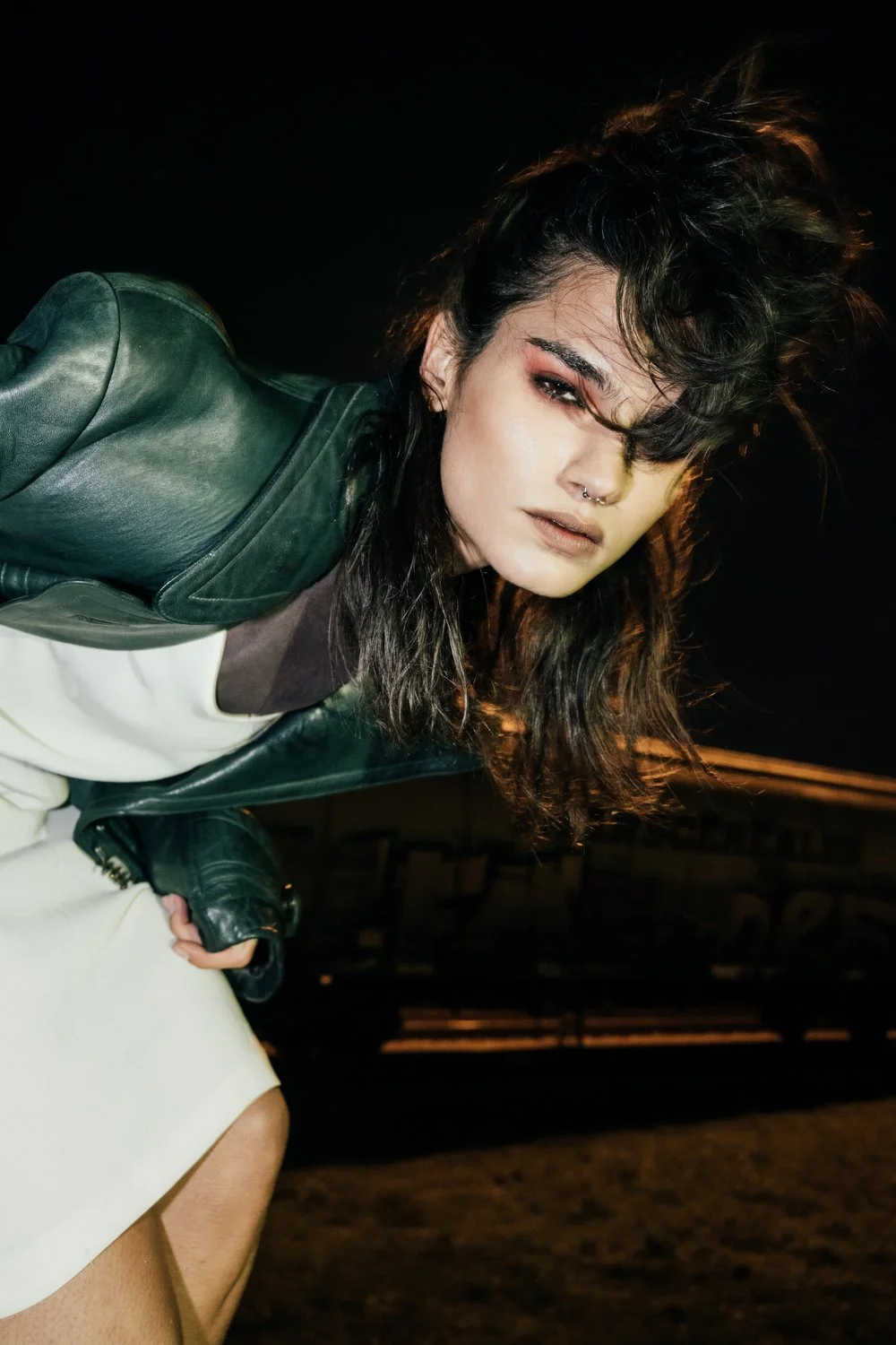 A young woman with dark, wavy hair and a septum piercing, wearing a dark leather jacket and posing at night.