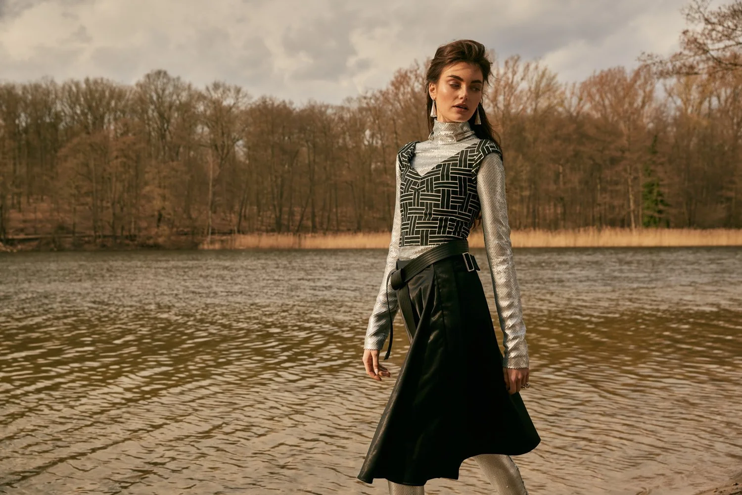 A young woman is standing by a river with a background of trees with autumn leaves, wearing a patterned top, a black skirt, and metallic boots, during cloudy weather.