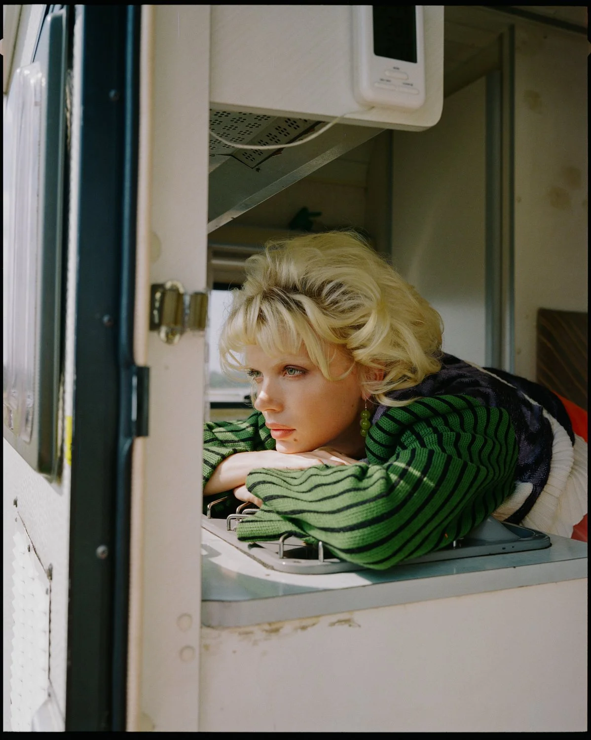 A woman with short blonde curly hair and green earrings is resting her chin on her folded arms, looking through an open refrigerator door.