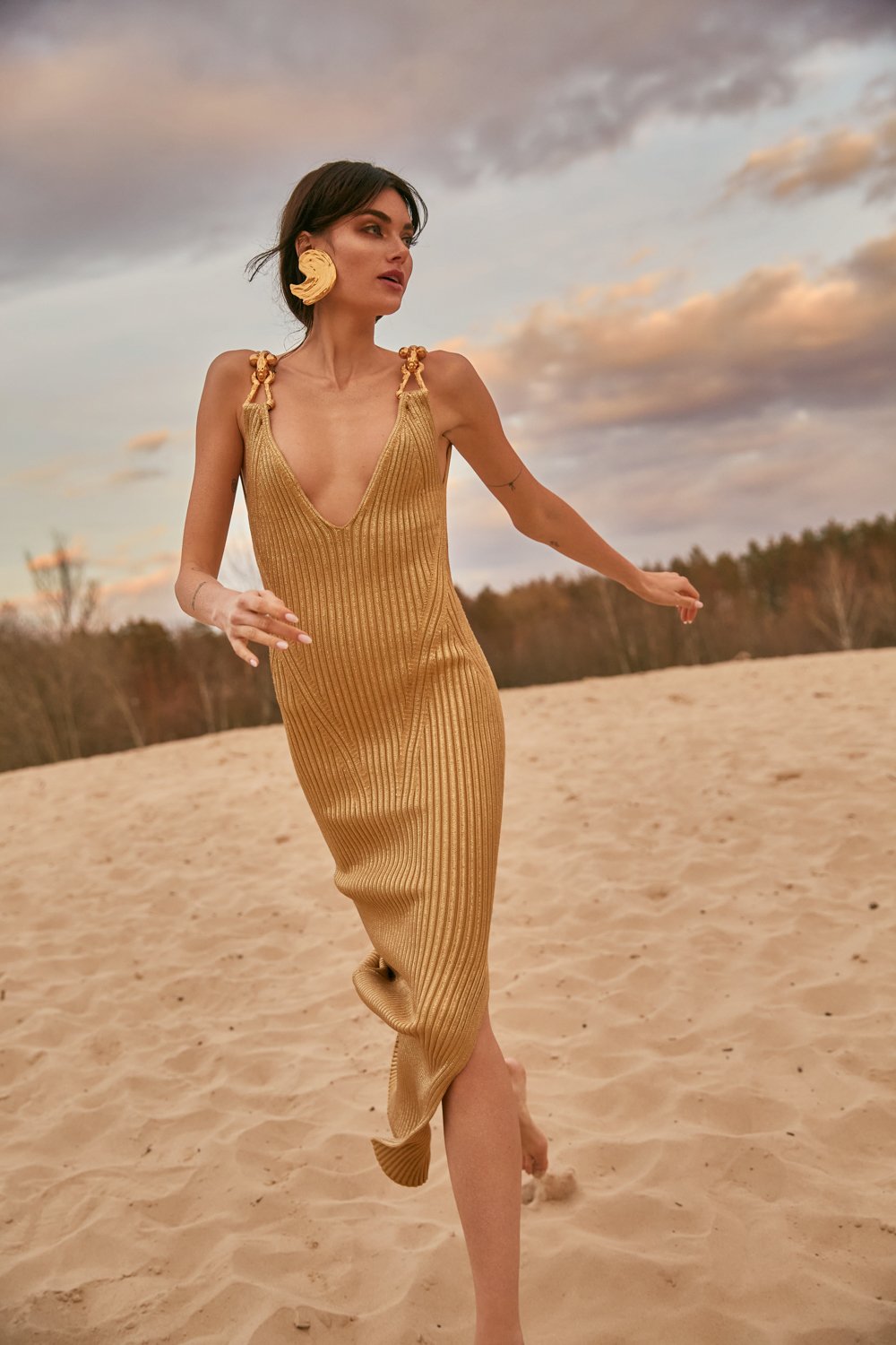 A woman with short dark hair wearing a gold, ribbed, sleeveless dress with tied shoulder straps, walking barefoot on a sandy beach during sunset, with cloudy sky and trees in the background.