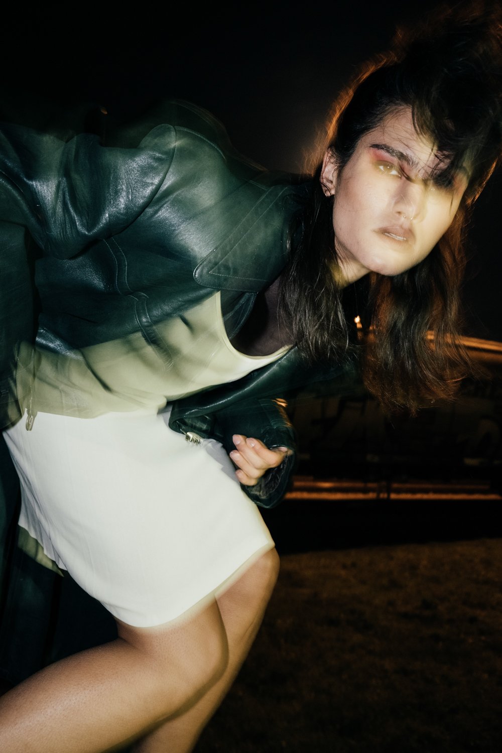 A woman with dark hair, wearing a black leather jacket, a beige top, and a white skirt, posing outdoors at night.