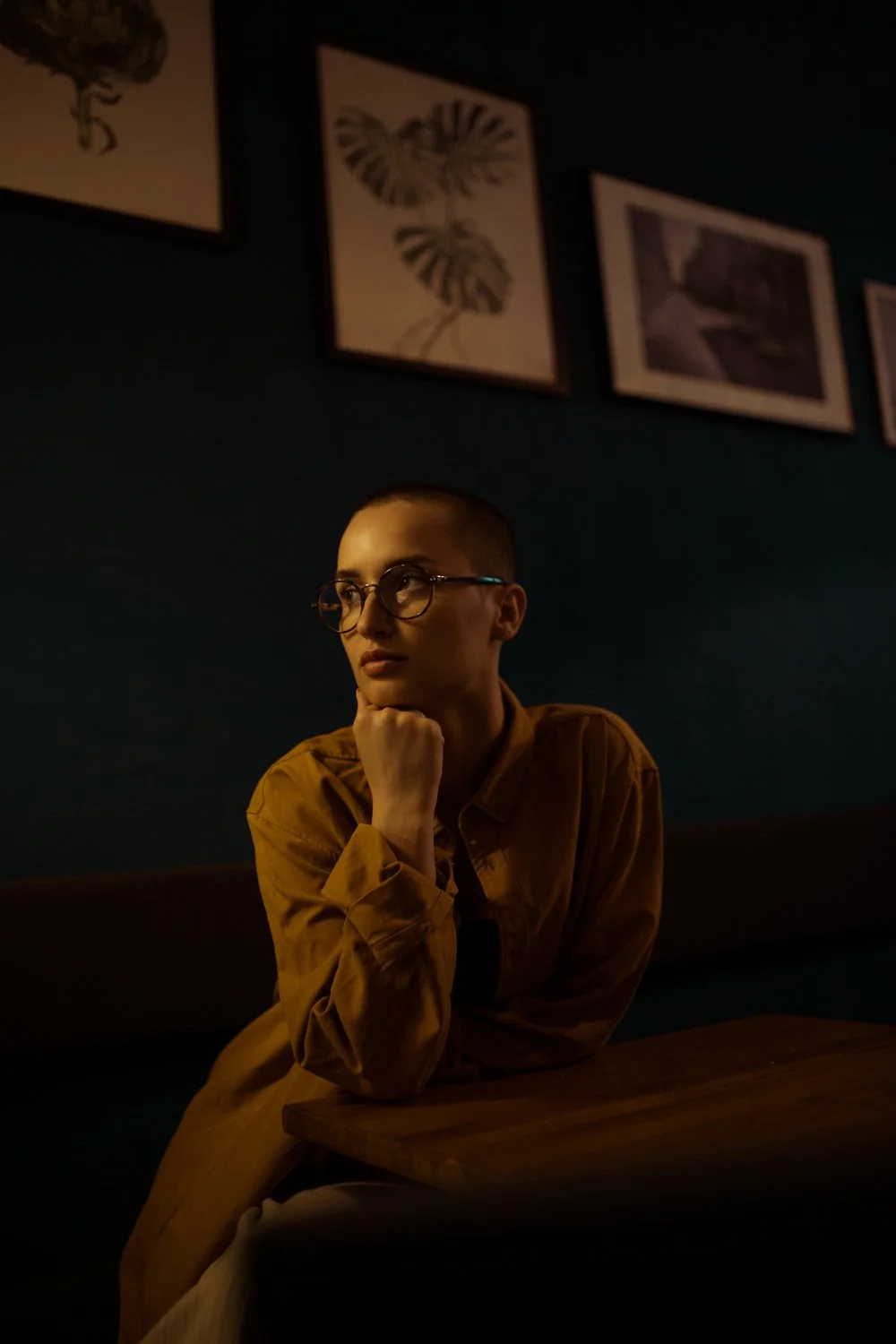 A woman with glasses and short hair wearing a mustard-colored jacket sitting at a dark wooden table in a dimly lit room with dark teal walls and framed artwork hanging above her.