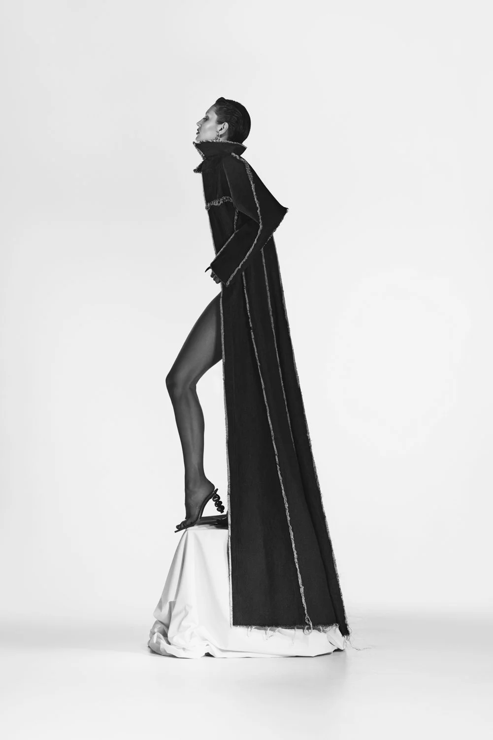 A model wearing a long, dark coat with embellishments, black hosiery, and high-heeled shoes, standing on a white platform against a plain white background.