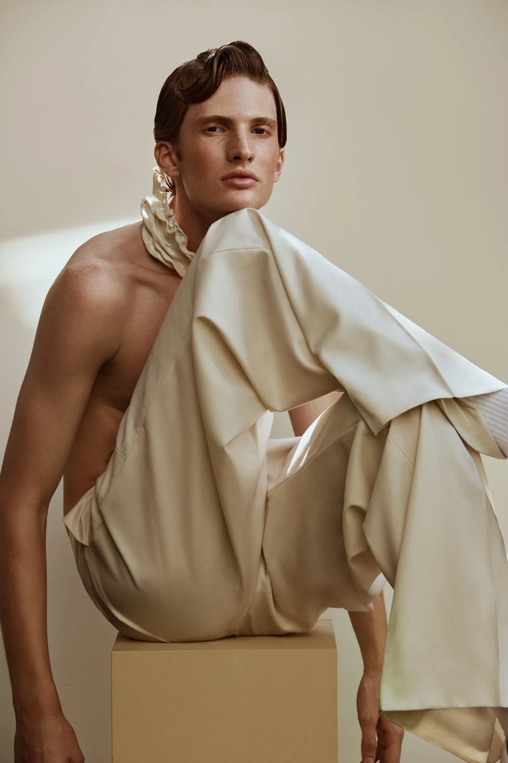 A shirtless young man with styled hair posing on a beige pedestal against a plain light background, wearing beige pants and a matching ruffled collar.