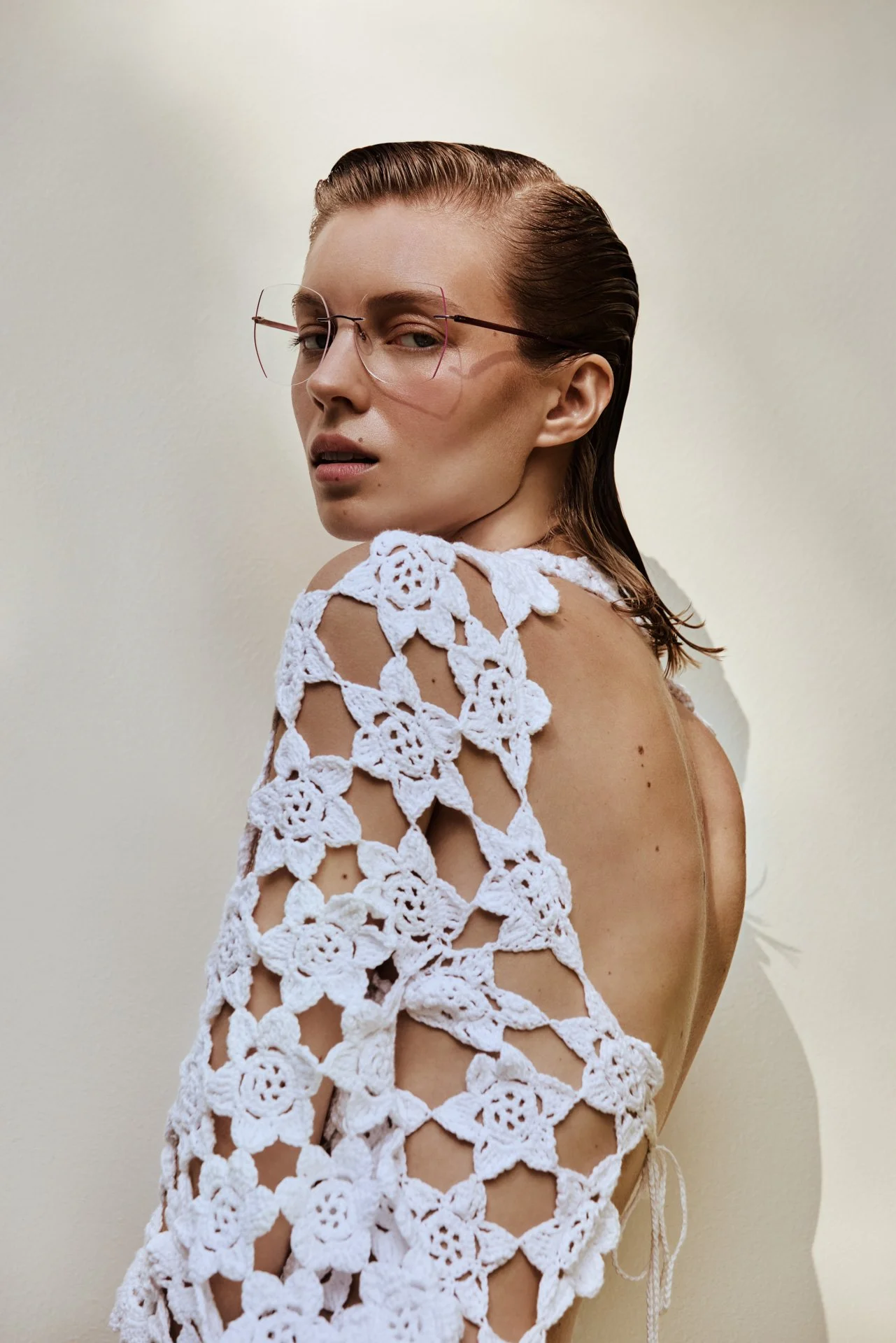 A woman with slicked-back hair, wearing glasses with a thin metal frame, and a white crocheted top with floral patterns, standing against a plain beige background.