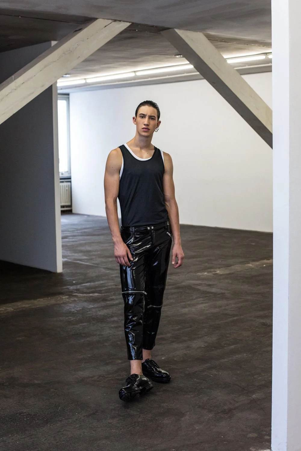 A young woman with dark hair pulled back, wearing a black sleeveless athletic top and shiny black pants, standing in an industrial-style interior space with white and dark walls and a concrete floor.