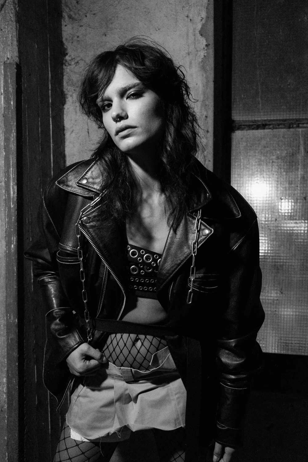 A woman with dark, wavy hair in a messy style, wearing a leather jacket over a fishnet top and shorts, posing in an industrial-style room with concrete walls and metal doors, in black and white.