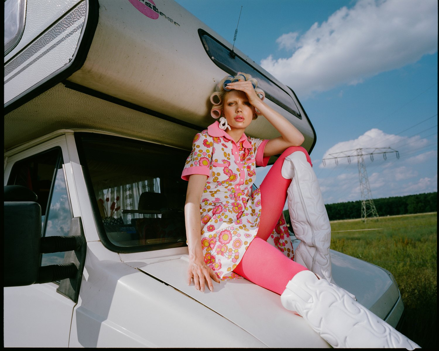 Woman in a colorful vintage dress with curlers in her hair, sitting on the hood of a white van in a rural field under a partly cloudy sky.