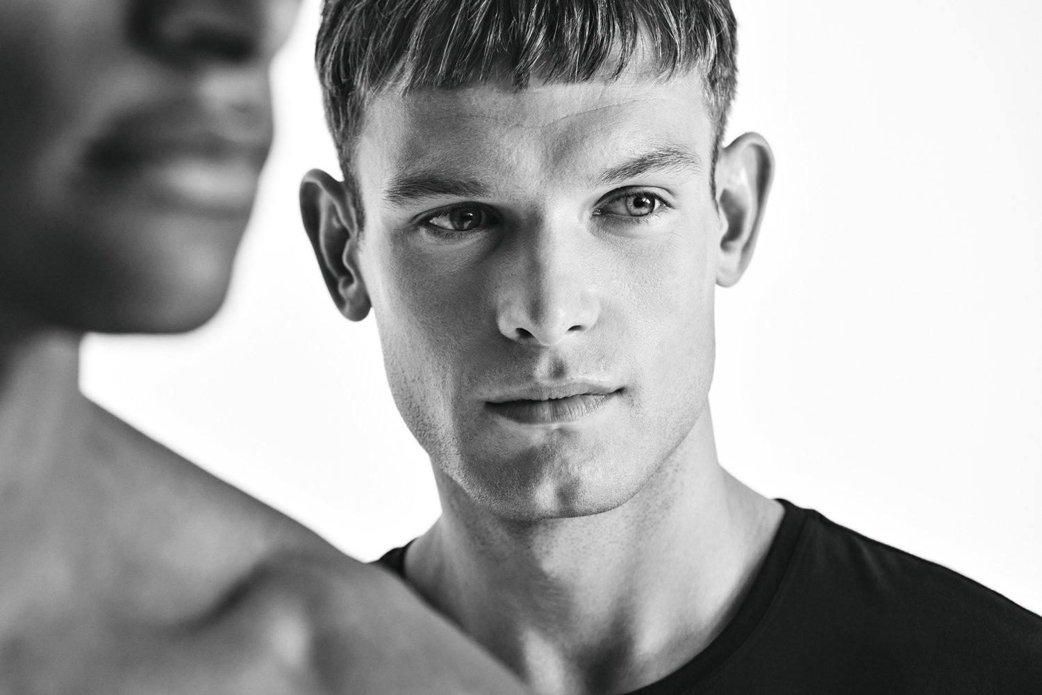 Close-up black and white photo of a young man with short hair, looking intently at a woman who is partially visible in the foreground.