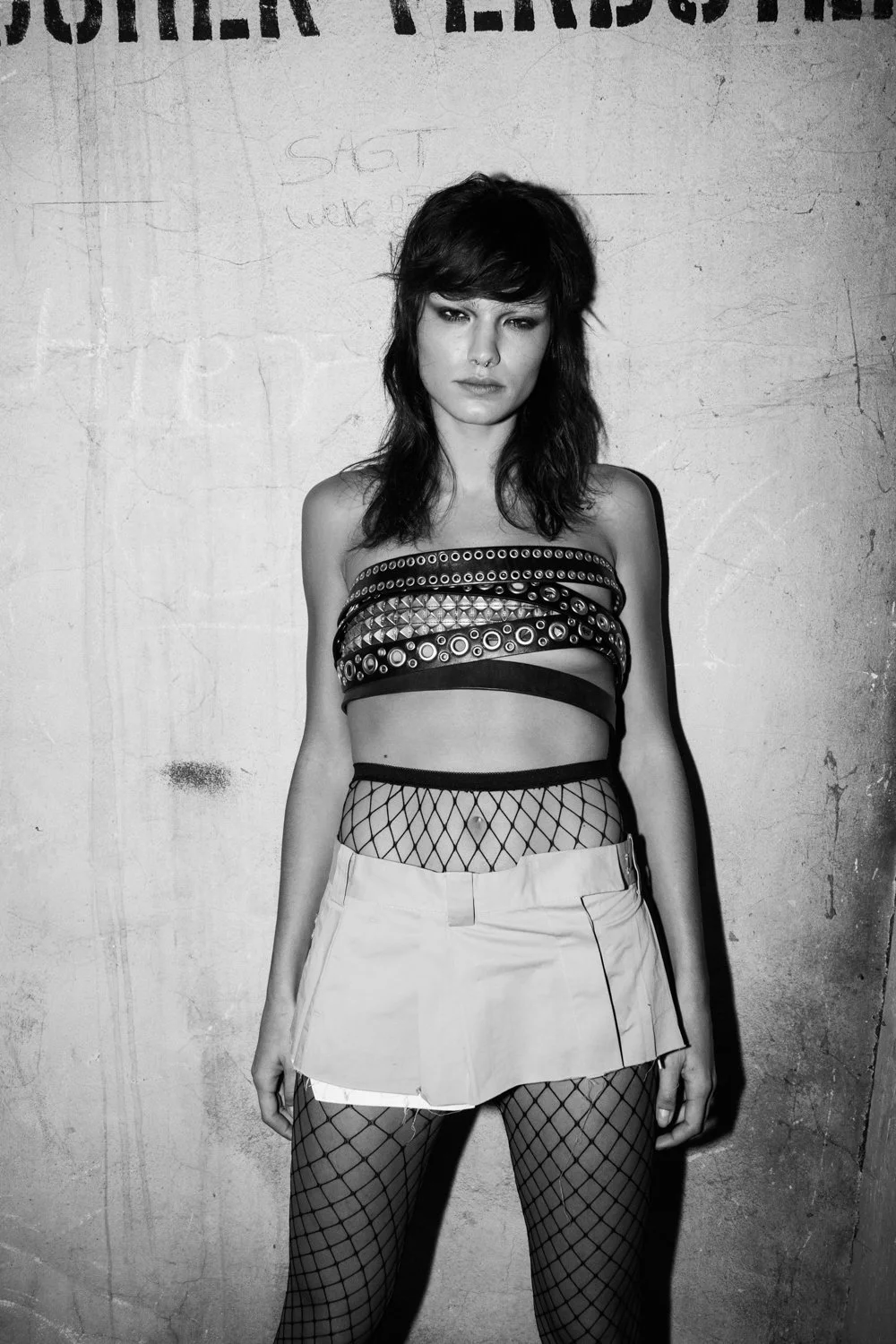 Black and white photo of a young woman with dark hair, wearing a strapless top with metal studs and rings, a short skirt, fishnet stockings, standing against a textured wall.
