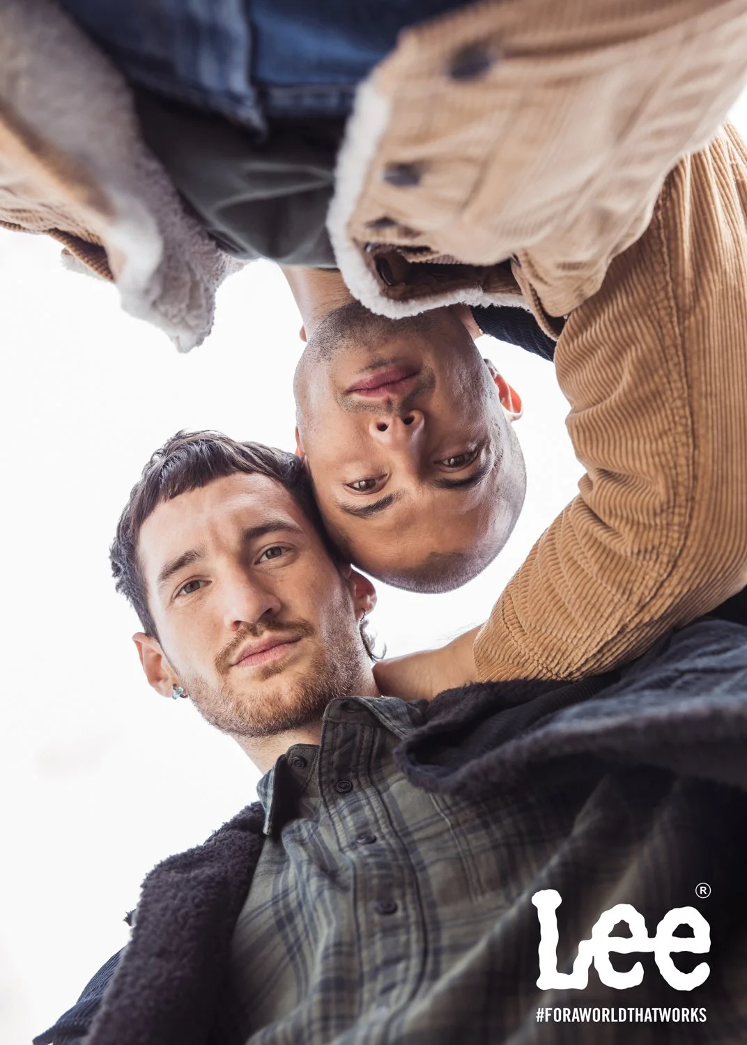 Two men lying on the ground looking into the camera, one with dark hair and beard, the other bald, close together, with a logo and hashtag at the bottom right corner.