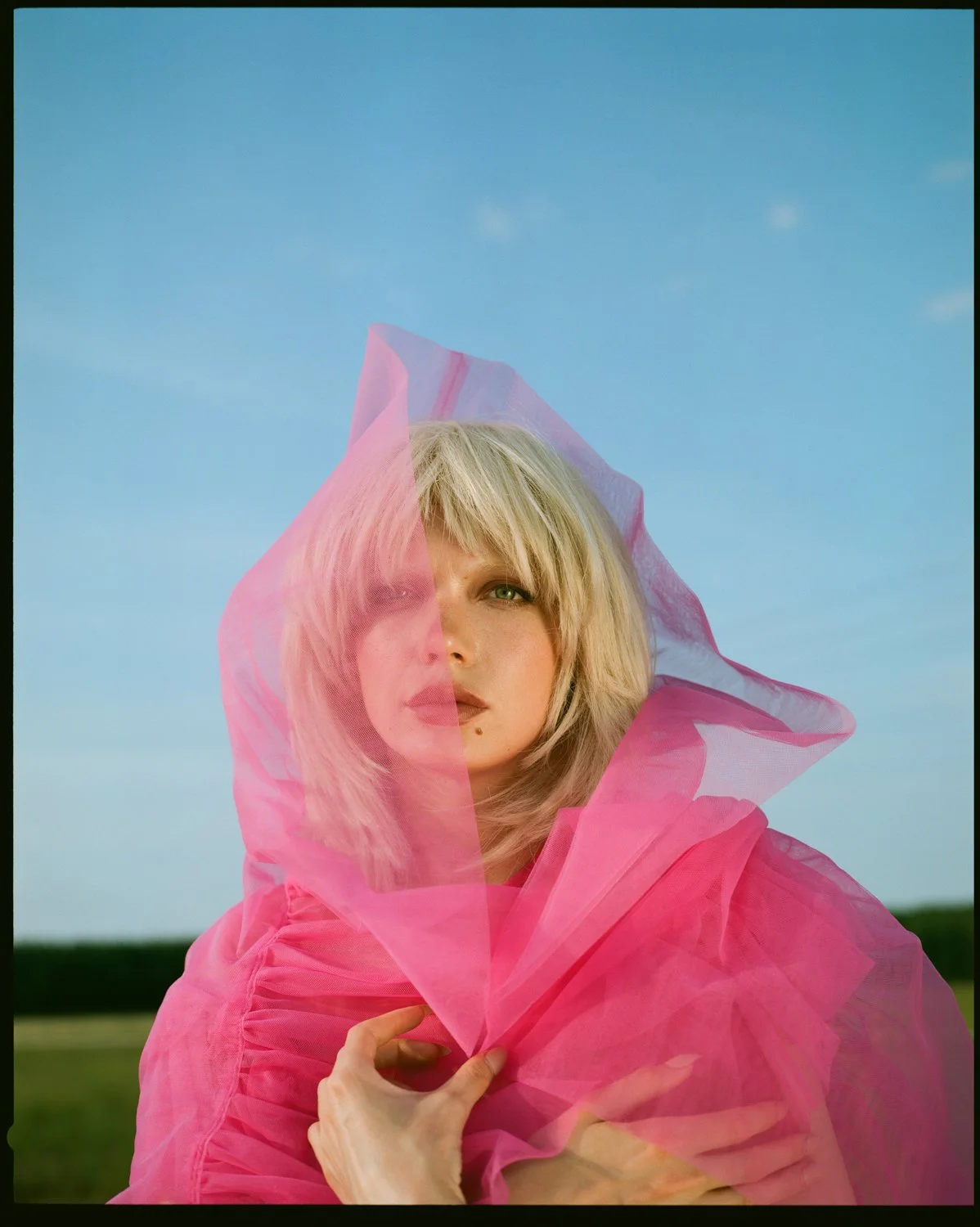 A woman with blonde hair and green eyes is holding a pink translucent fabric near her face outdoors against a background of blue sky and green fields.