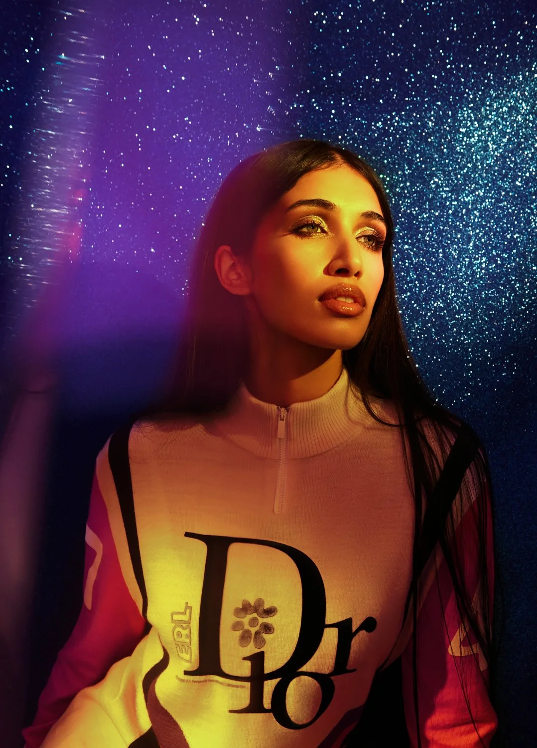 A young woman with long dark hair and bright makeup gazes to the side against a glittery blue background with colorful lighting.