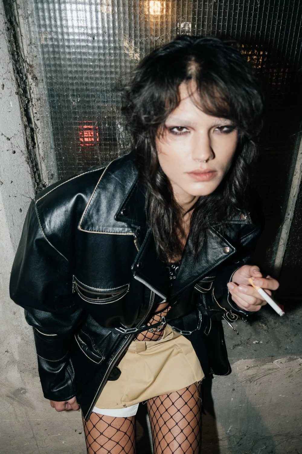 A woman with dark, wavy hair and a piercing, wearing a black leather jacket, fishnet stockings, and holding a cigarette, leaning against a gritty, textured wall with a metal grid behind her.