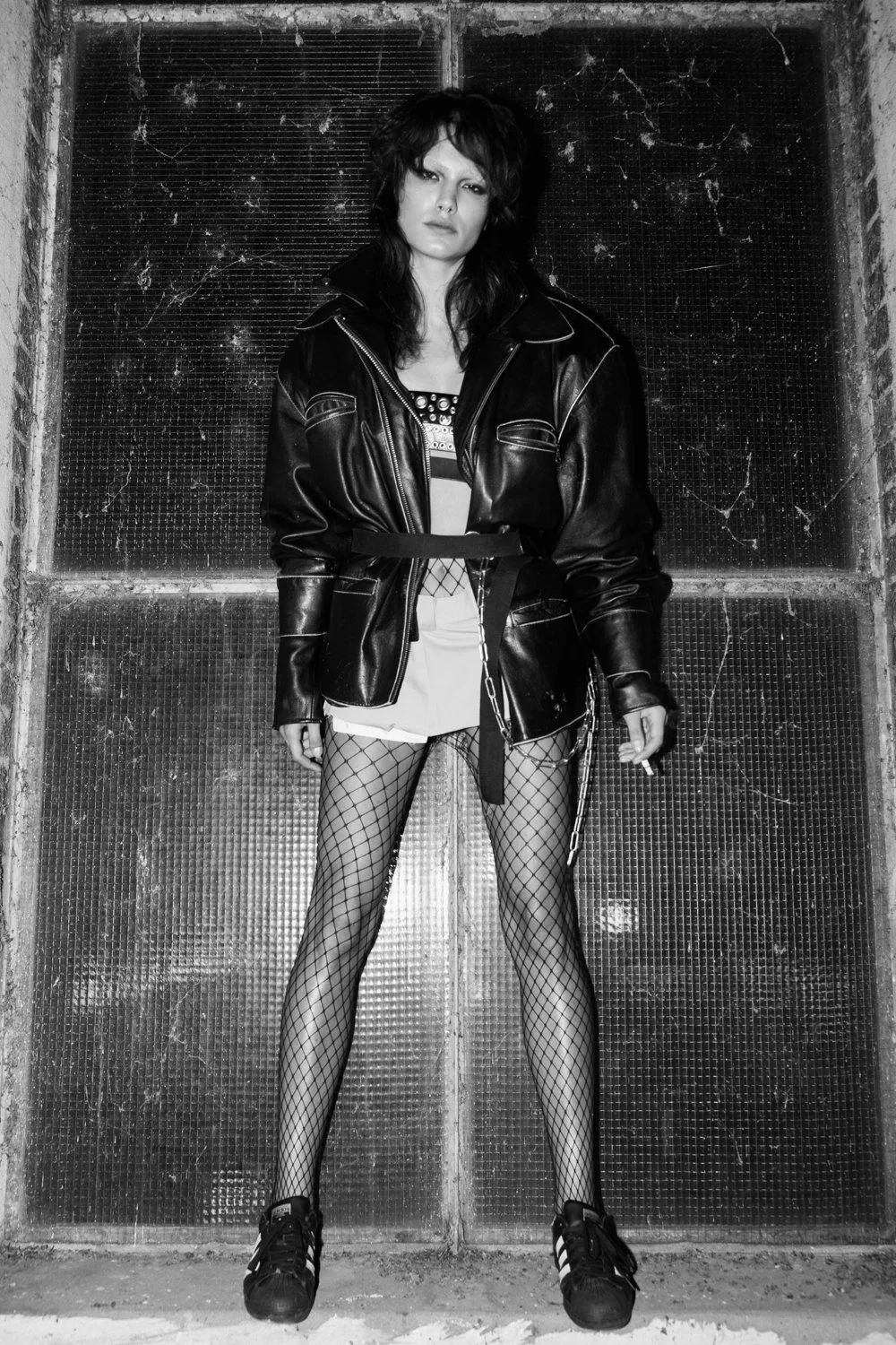 A woman standing against a textured wall, wearing a black leather jacket, fishnet stockings, sneakers, and a revealing outfit underneath.
