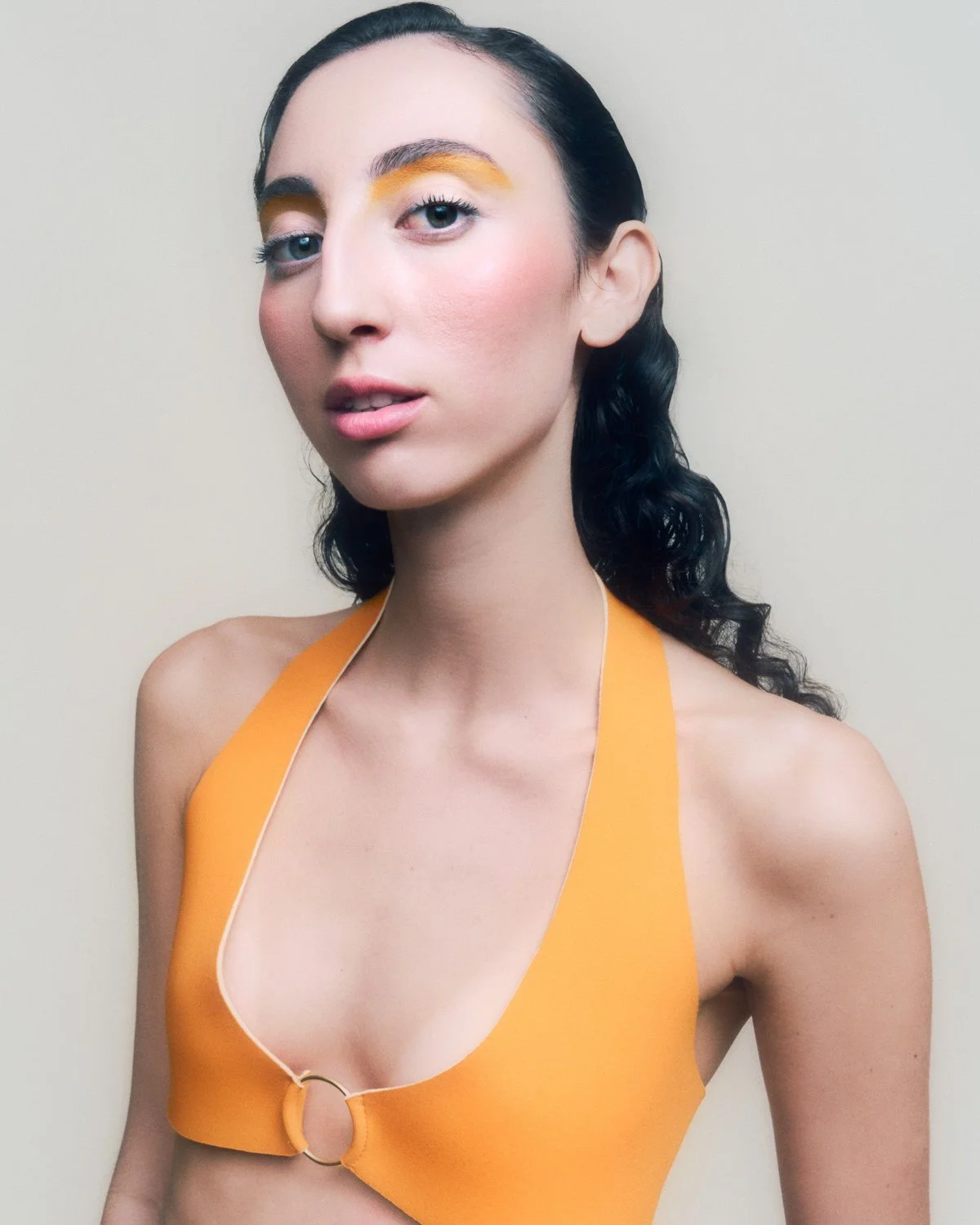 A woman with dark wavy hair and blue eyes wearing minimal makeup, a yellow sleeveless top with a halter neck and ring detail, standing against a plain light background.