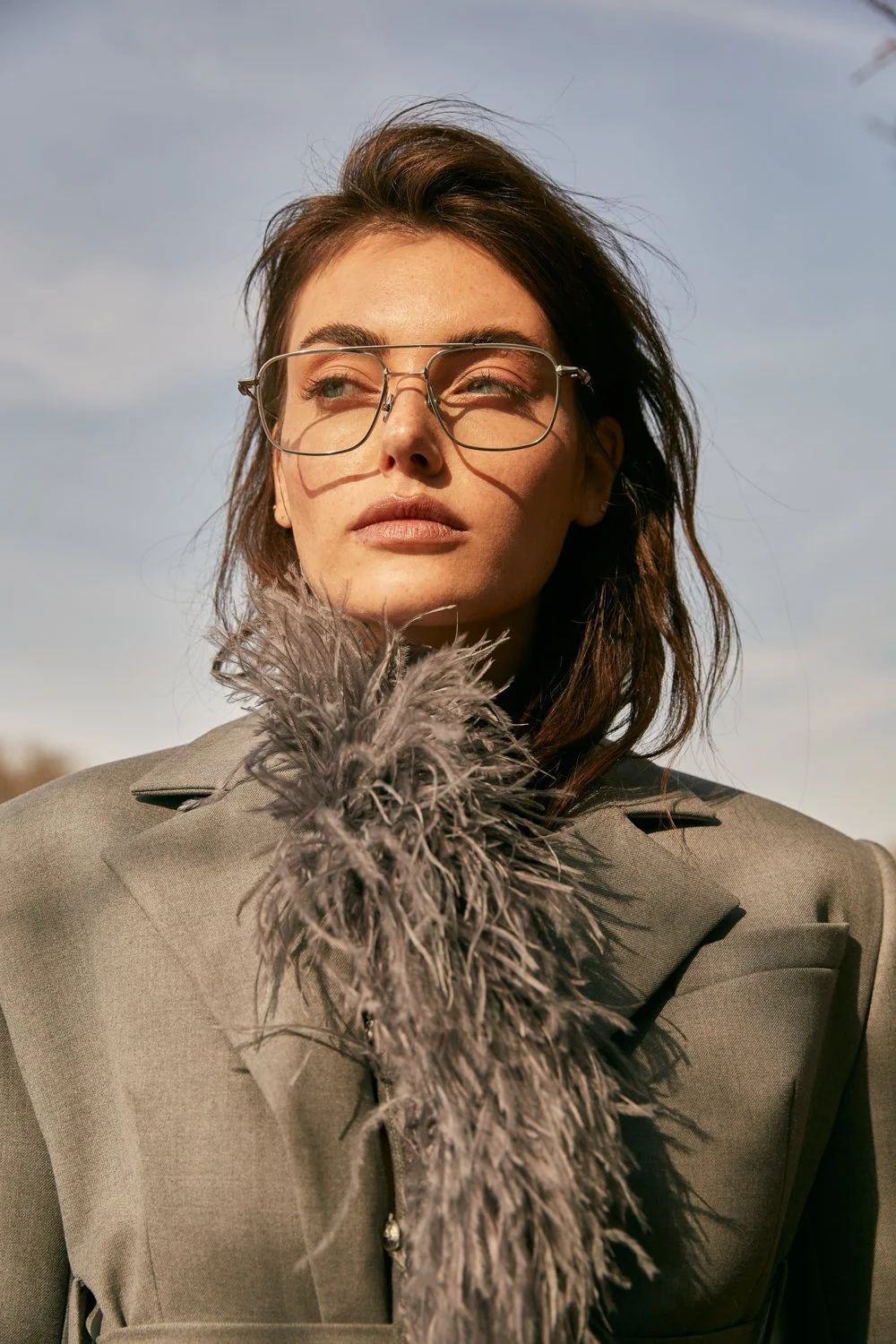 A young woman with brown hair and glasses looking into the distance outdoors, wearing a beige coat with a feathered collar.