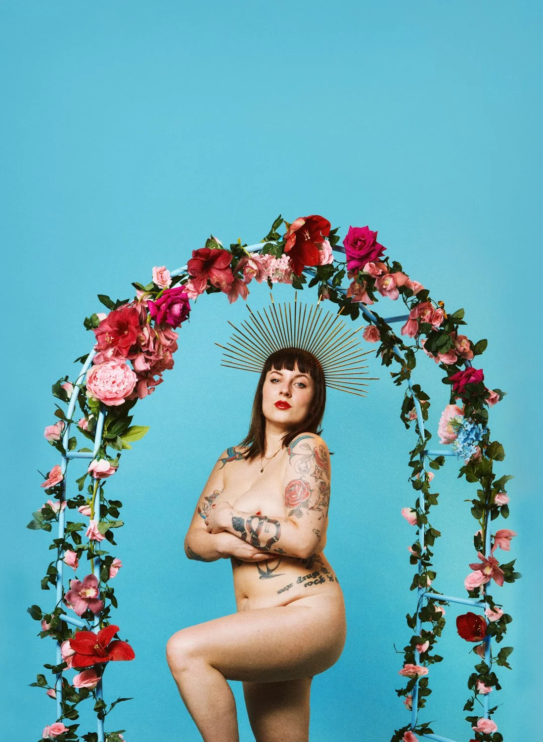 A nude woman with tattoos on her arms and legs, standing inside a floral arch with pink and red flowers, on a blue background, wearing a metallic halo-like headpiece.