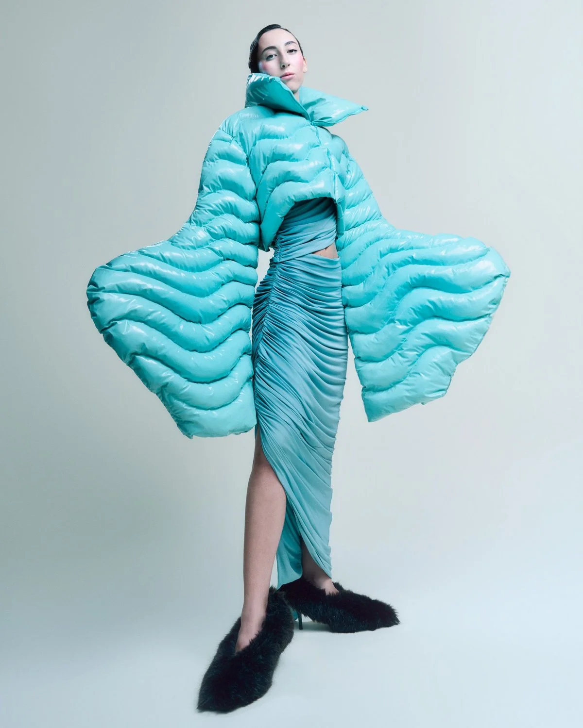 A woman in a surreal blue puffer jacket with large, curving sleeves and a matching ruched dress with a high slit, wearing black fuzzy high heels, standing against a plain light background.