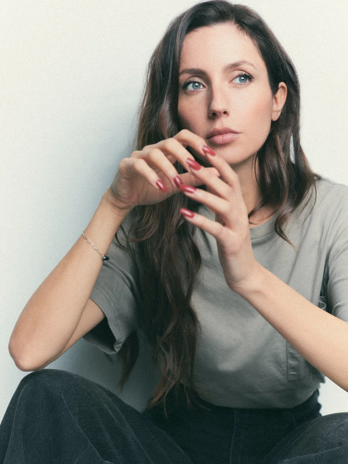 A woman with long brown hair and blue eyes, wearing a gray t-shirt, sitting against a plain white background.