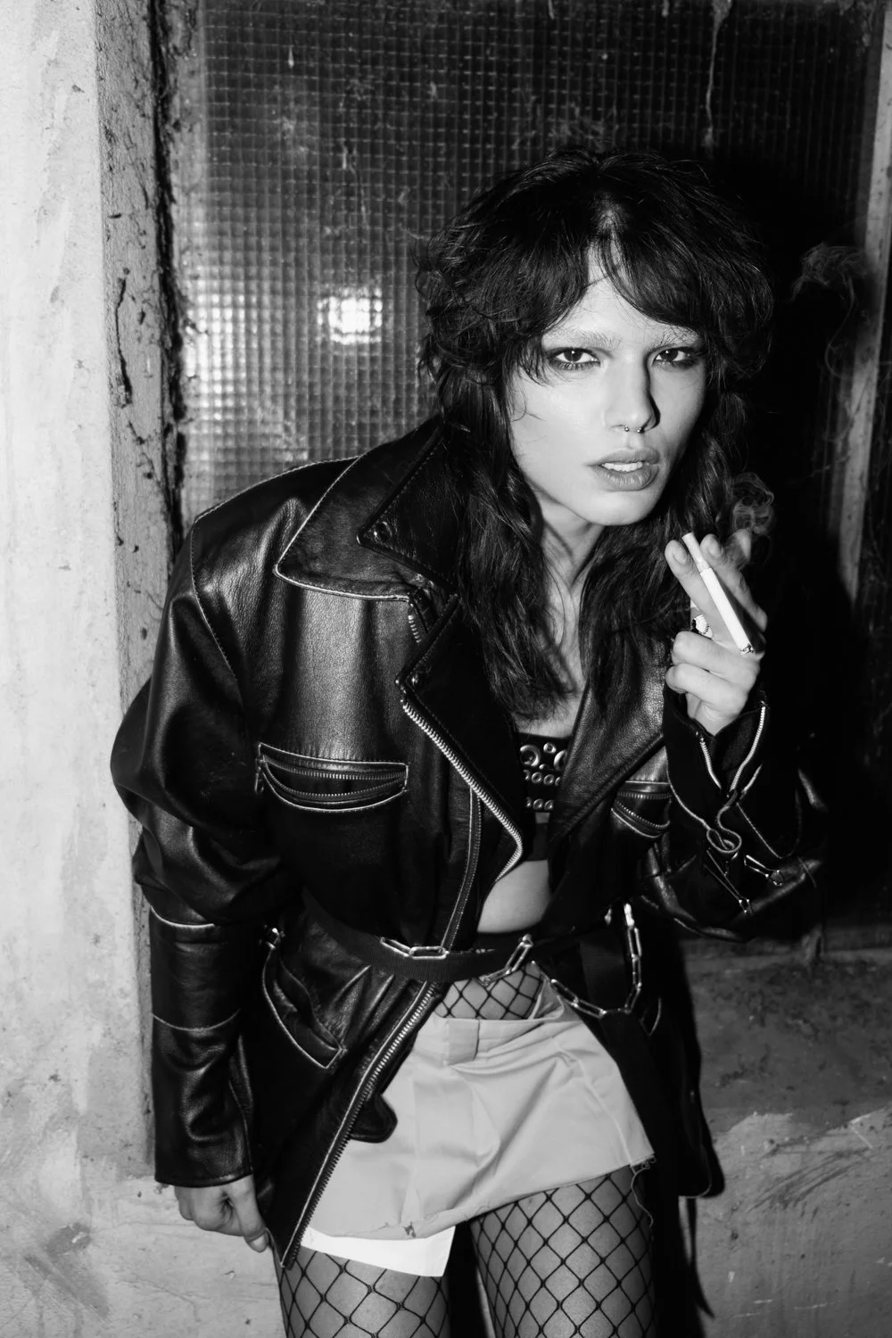 Black and white photo of a young woman with dark, wavy hair, wearing a leather jacket, fishnet stockings, and a skirt, holding a cigarette, standing in front of a textured window.