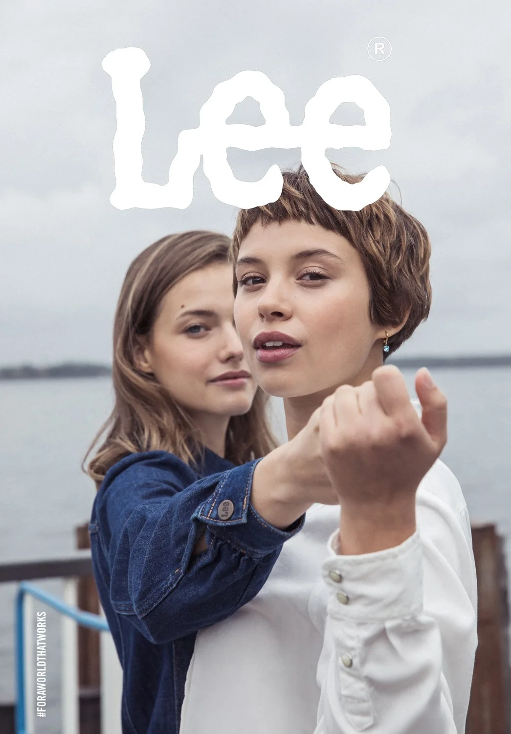 Two women standing outdoors near a body of water under an overcast sky, with one woman in white and the other in a denim jacket, holding her fist towards the camera, with the word 'Lee' in white text overlay.