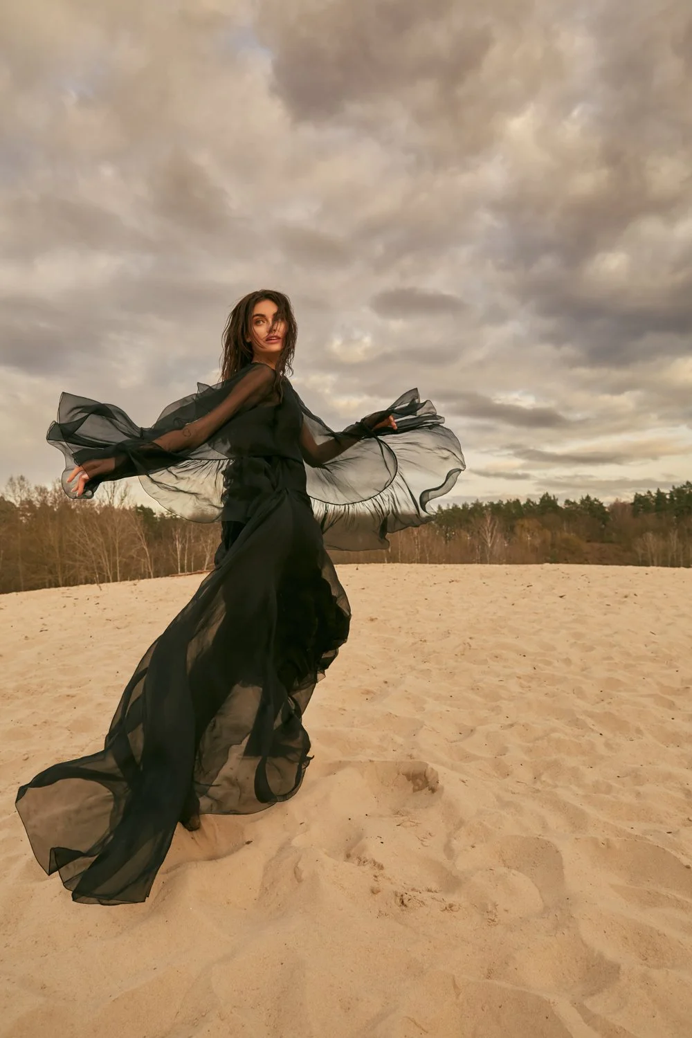 A woman in a flowing black dress standing on a sandy beach with cloudy skies in the background.